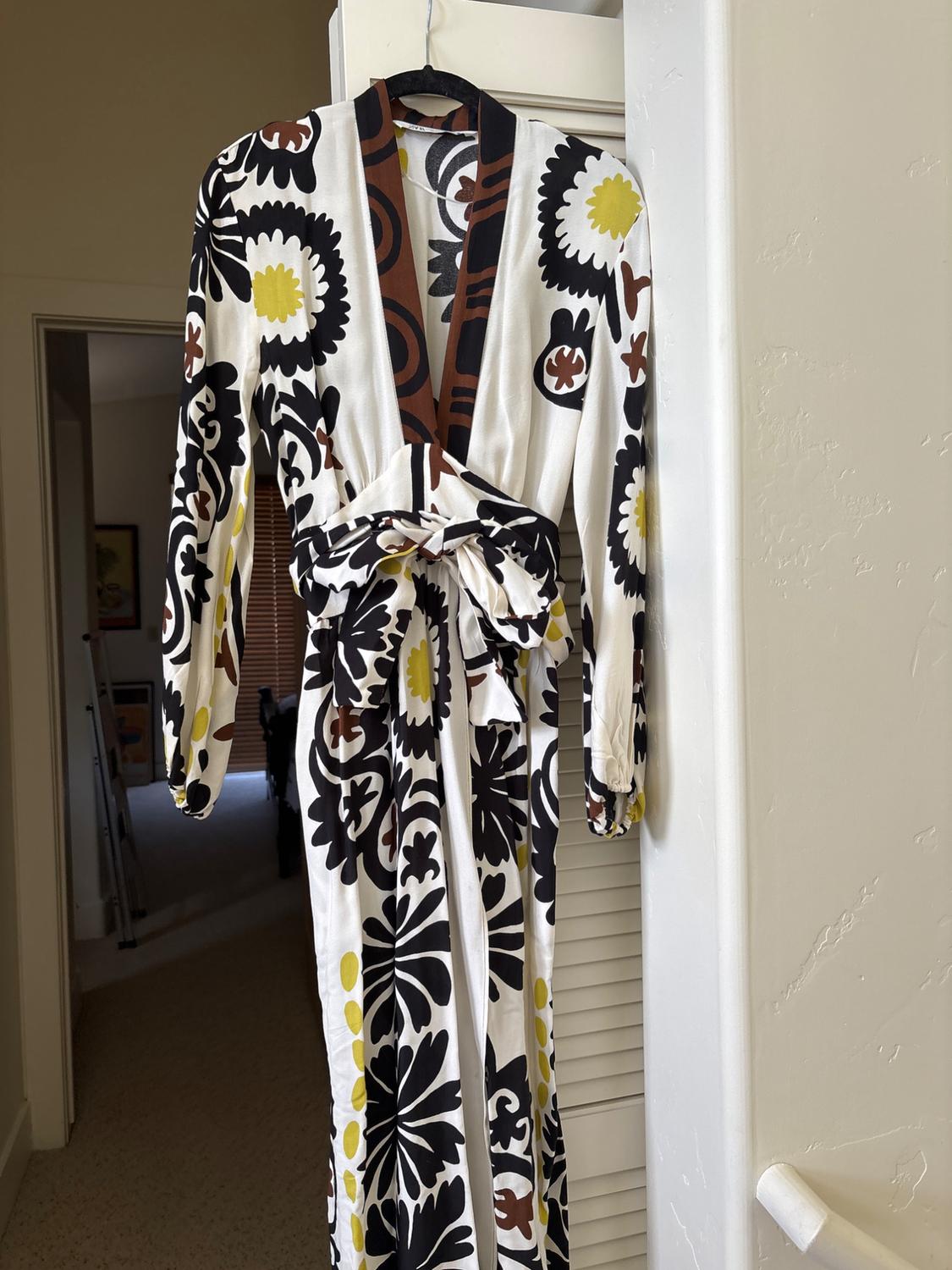 White maxi dress with a large black, brown, and yellow floral print. It features a V-neckline and a tie waist, hanging on a door at home.