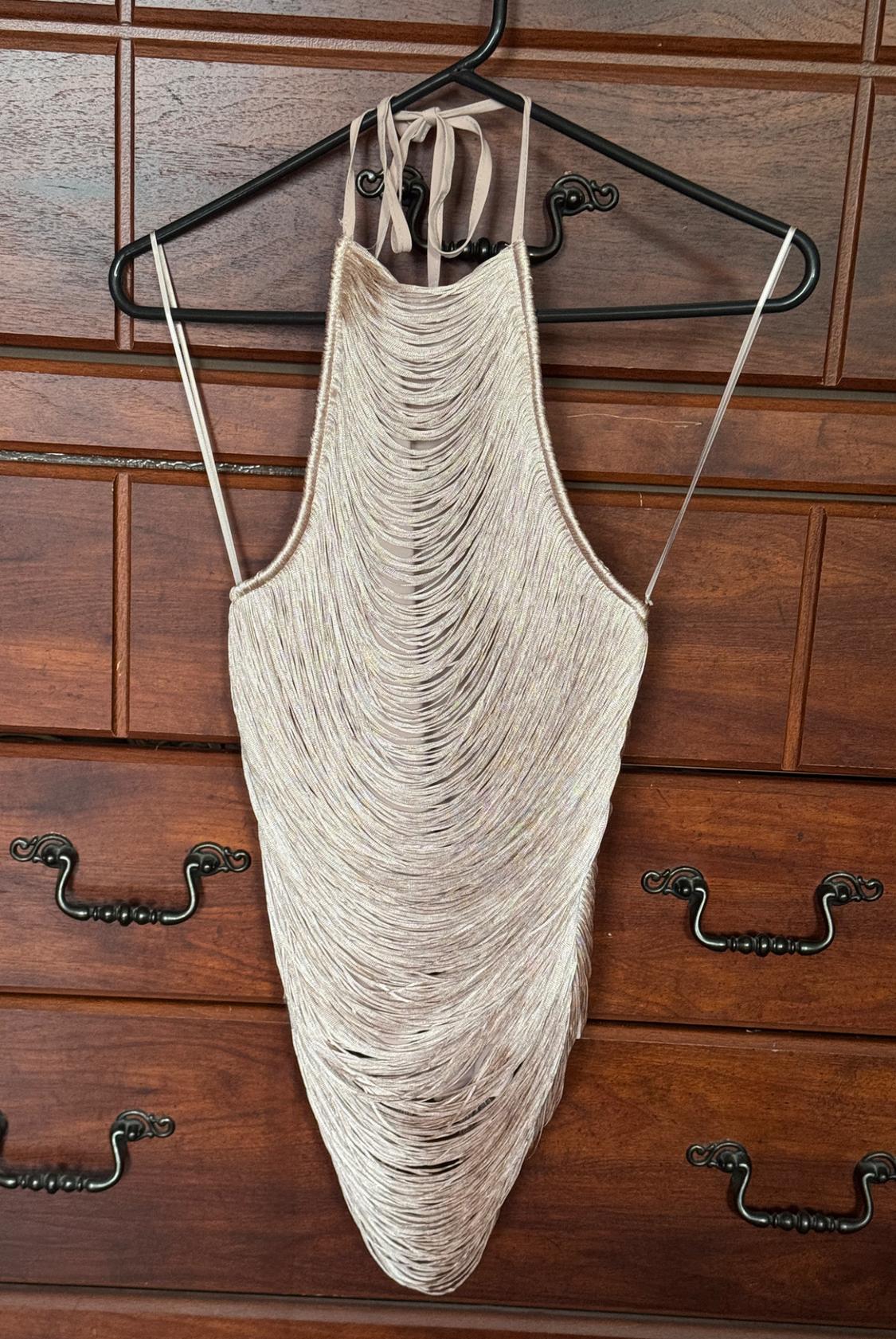 Cream-colored halter top made of cascading fringe, hanging on a black hanger in front of the drawers of a dark wood dresser.