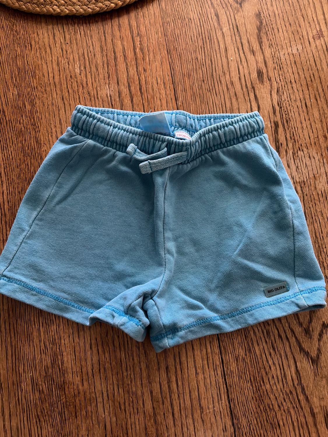 Blue shorts with an elasticated drawstring waist, on a wooden table. Visible tag: "BIG OLITA".