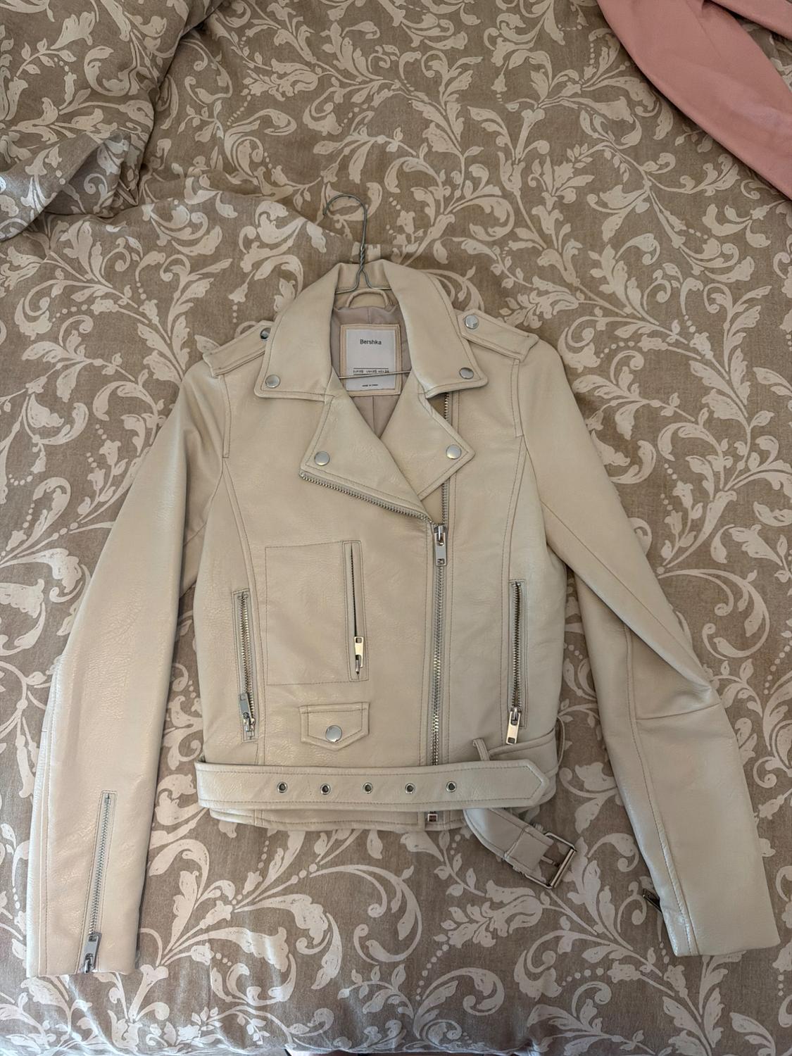 Beige biker jacket with zips and a belt, on a hanger above a bed with a floral-patterned bedspread. Text: Bershka.