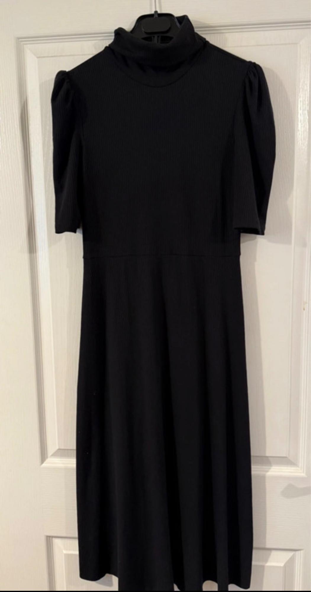 Black short-sleeved high-neck dress, hanging on a white door. Simple and elegant design.