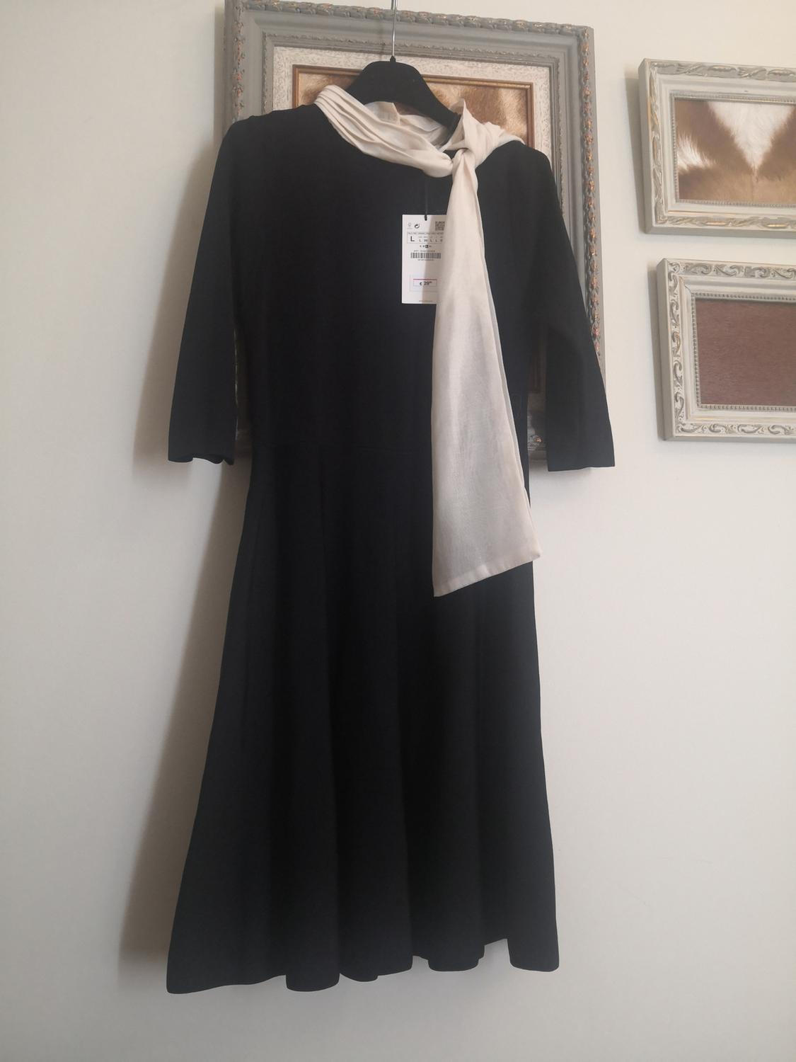 Black three-quarter sleeve dress hanging on a hanger, accompanied by a white scarf. Background with decorative frames on the wall.