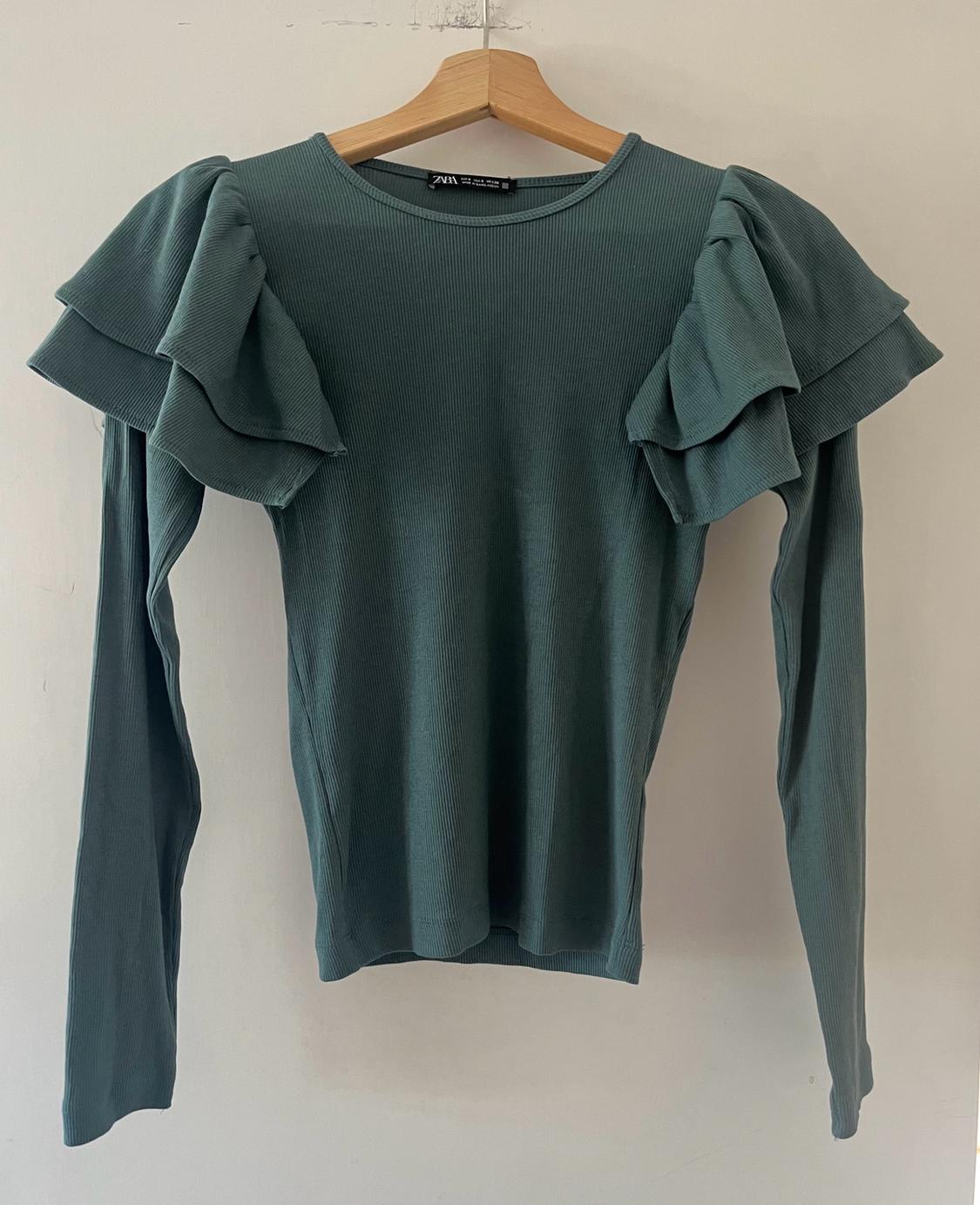 Green ribbed long-sleeved T-shirt with frills on the shoulders, hanging on a wooden hanger against a white background. Text ZARA.