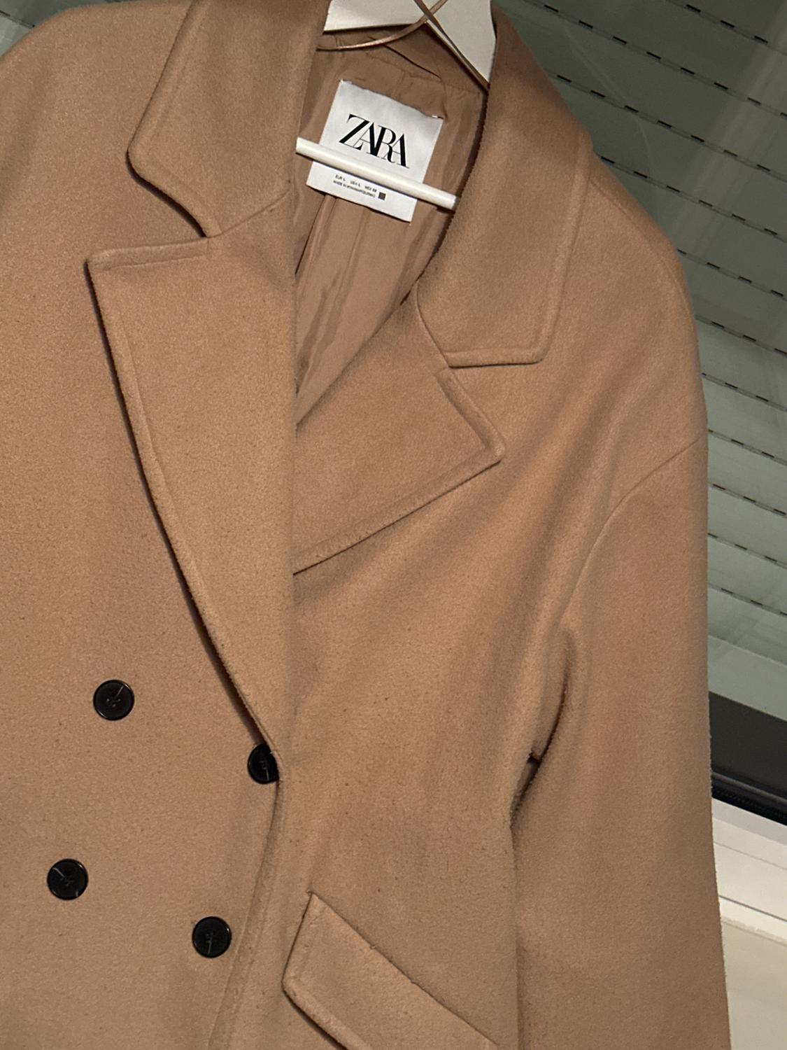 Camel-coloured double-breasted coat with black buttons hanging on a white hanger. Label with the text ZARA. Interior background with white Venetian blind.