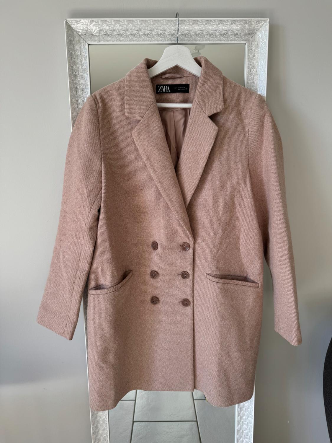 Light pink double-breasted coat hanging on a white hanger in front of a mirror with a silver frame. Label visible.