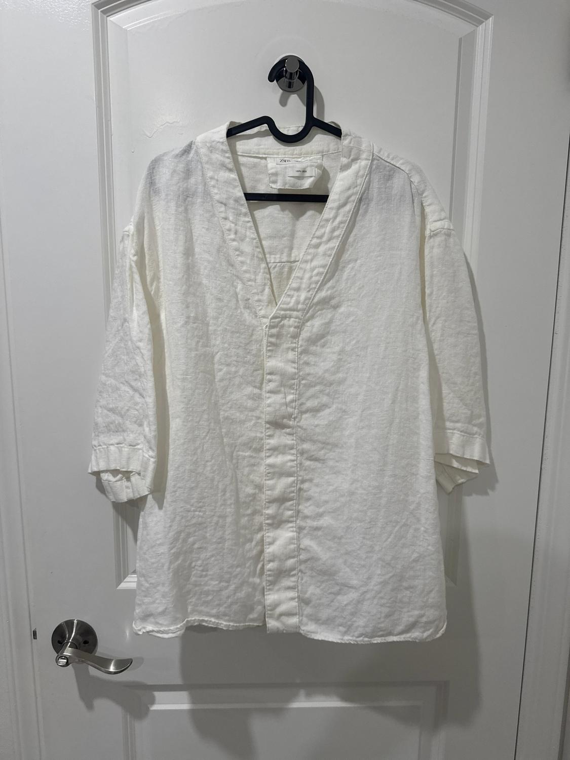 Loose white shirt with a V-neck and short sleeves, hanging on a white door. Tag with the text ZARA 100% LINEN.