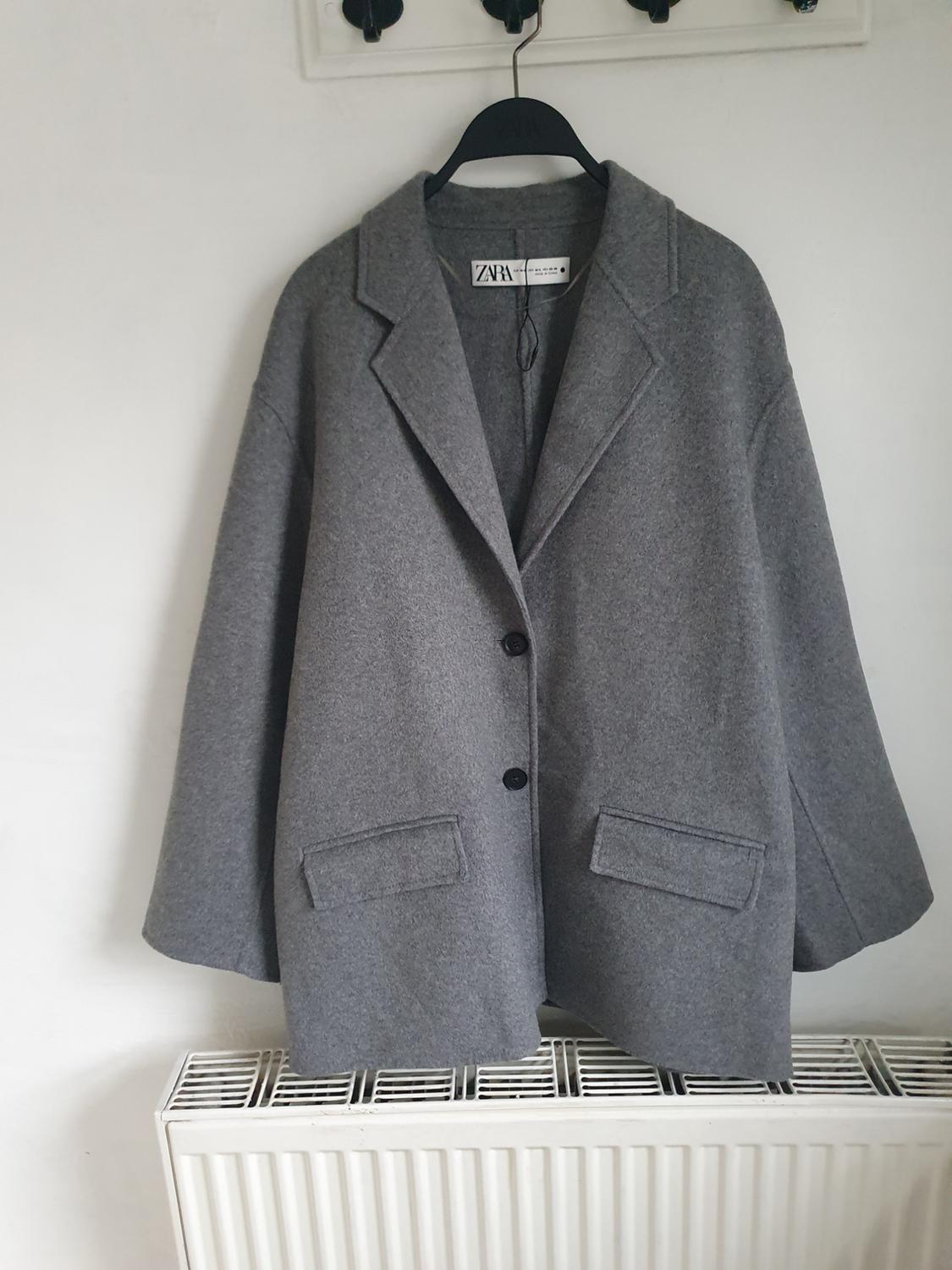 Grey wool coat hanging on a black hanger over a white radiator. Visible label with the text "ZARA".