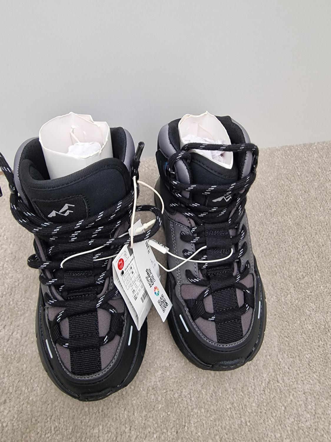 Black and grey boots with laces, hanging tags, and paper stuffing inside, on a beige carpet.