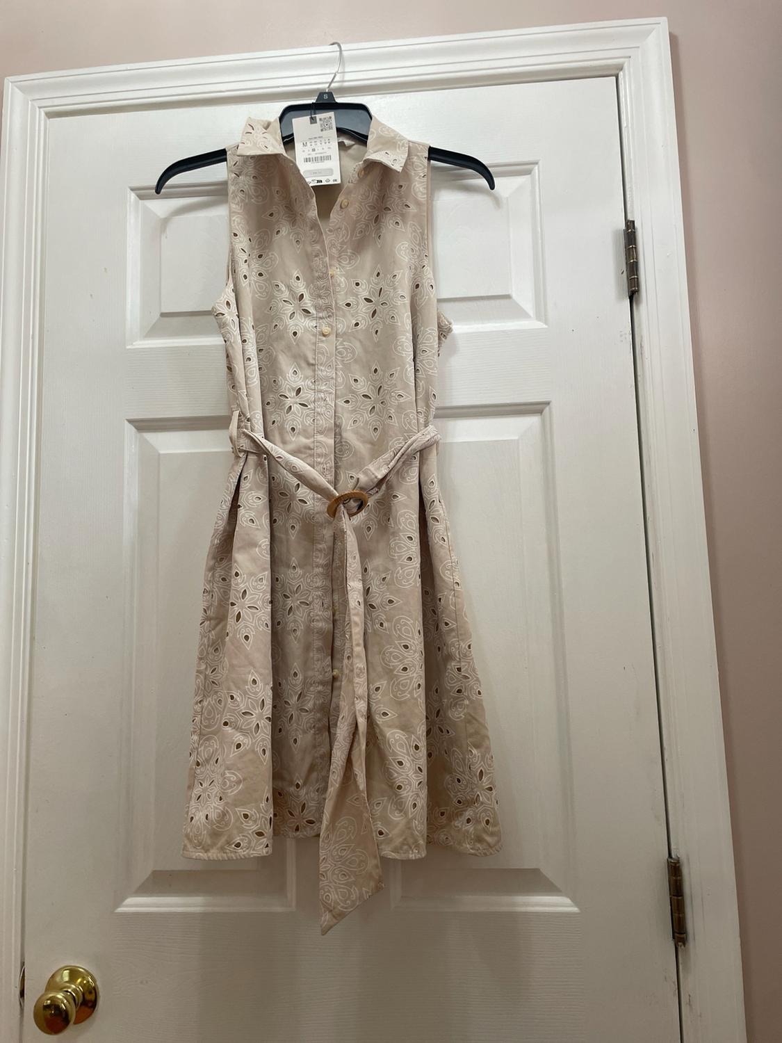 Sleeveless beige shirt dress with cutout embroidery and a belt, hanging on a hanger over a white door.
