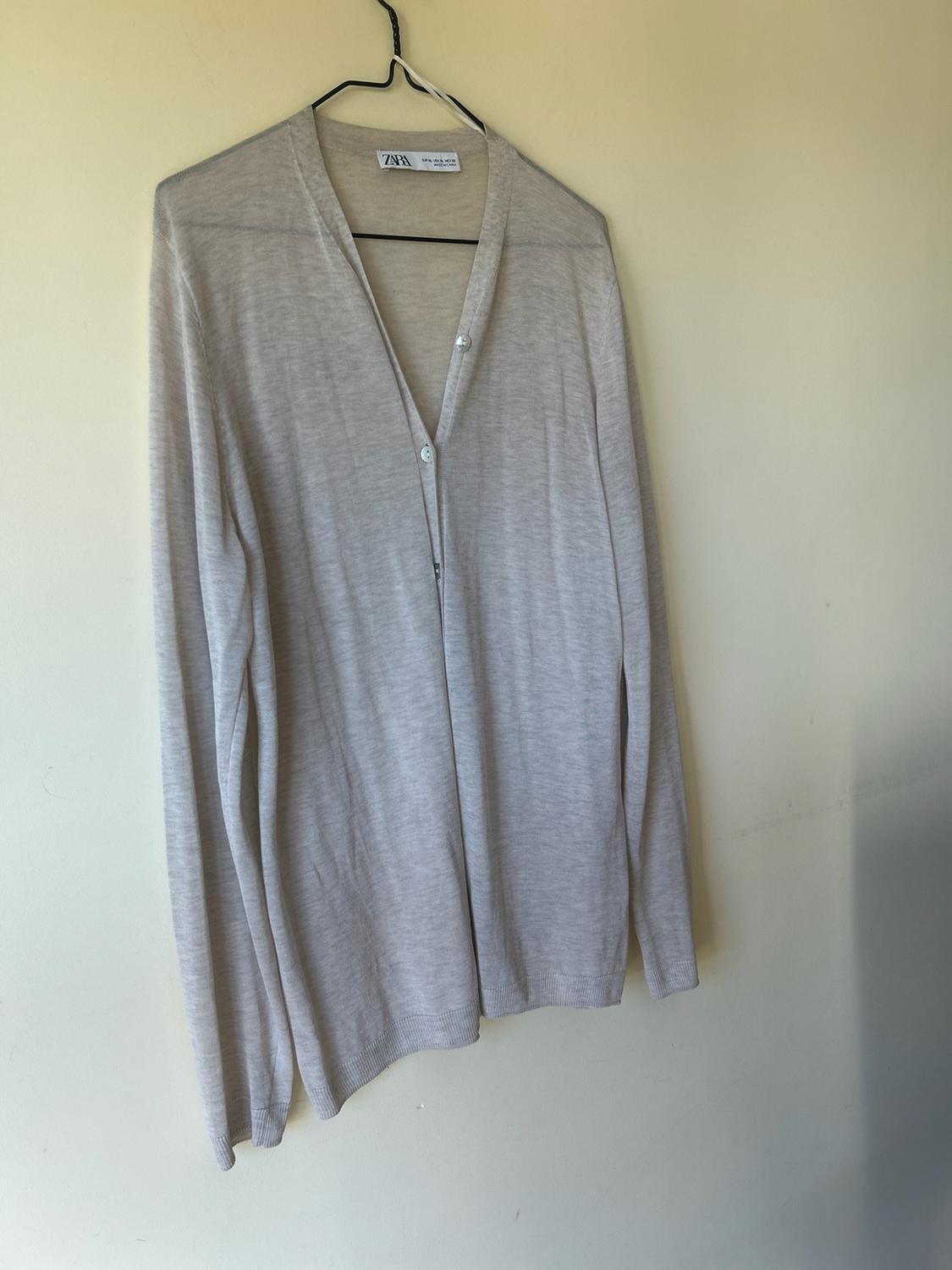 Fine beige long-sleeved cardigan with a V-neck and buttons, hanging on a hanger against a light-coloured wall. ZARA label.