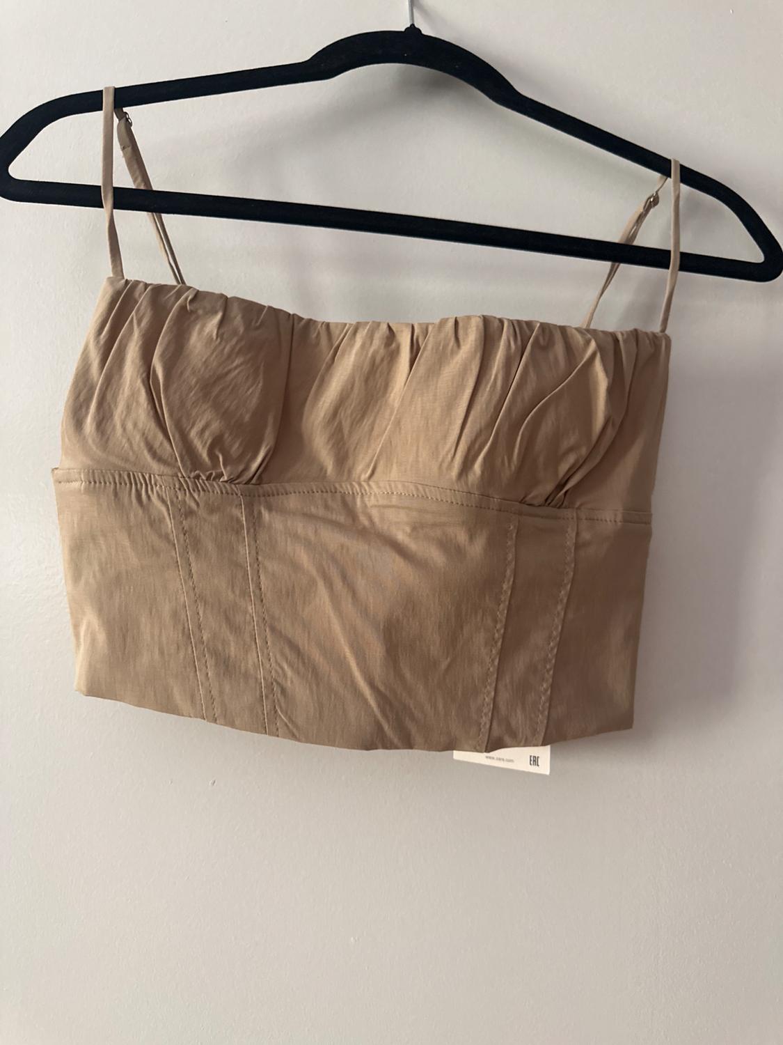 Beige shirred camisole crop top, hanging on a black hanger against a white wall. Label with text: www.zara.com EAC.