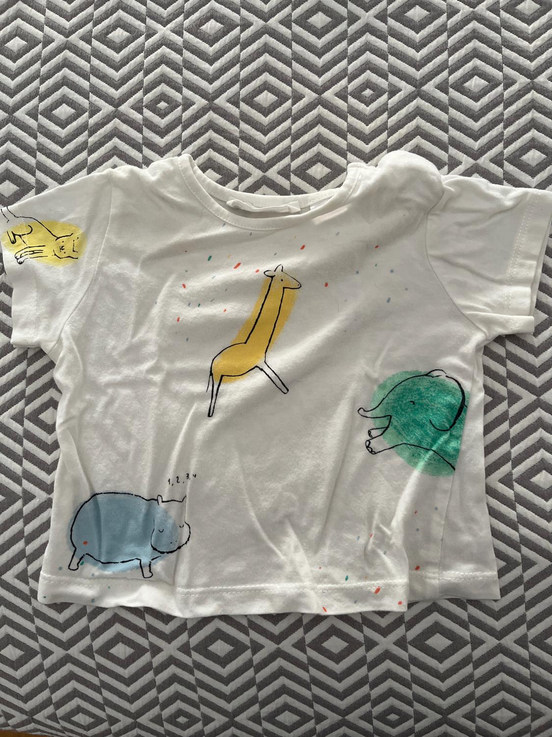 Children's white t-shirt with animal drawings: yellow giraffe, blue hippopotamus, green elephant, and yellow lion on a grey geometric background.