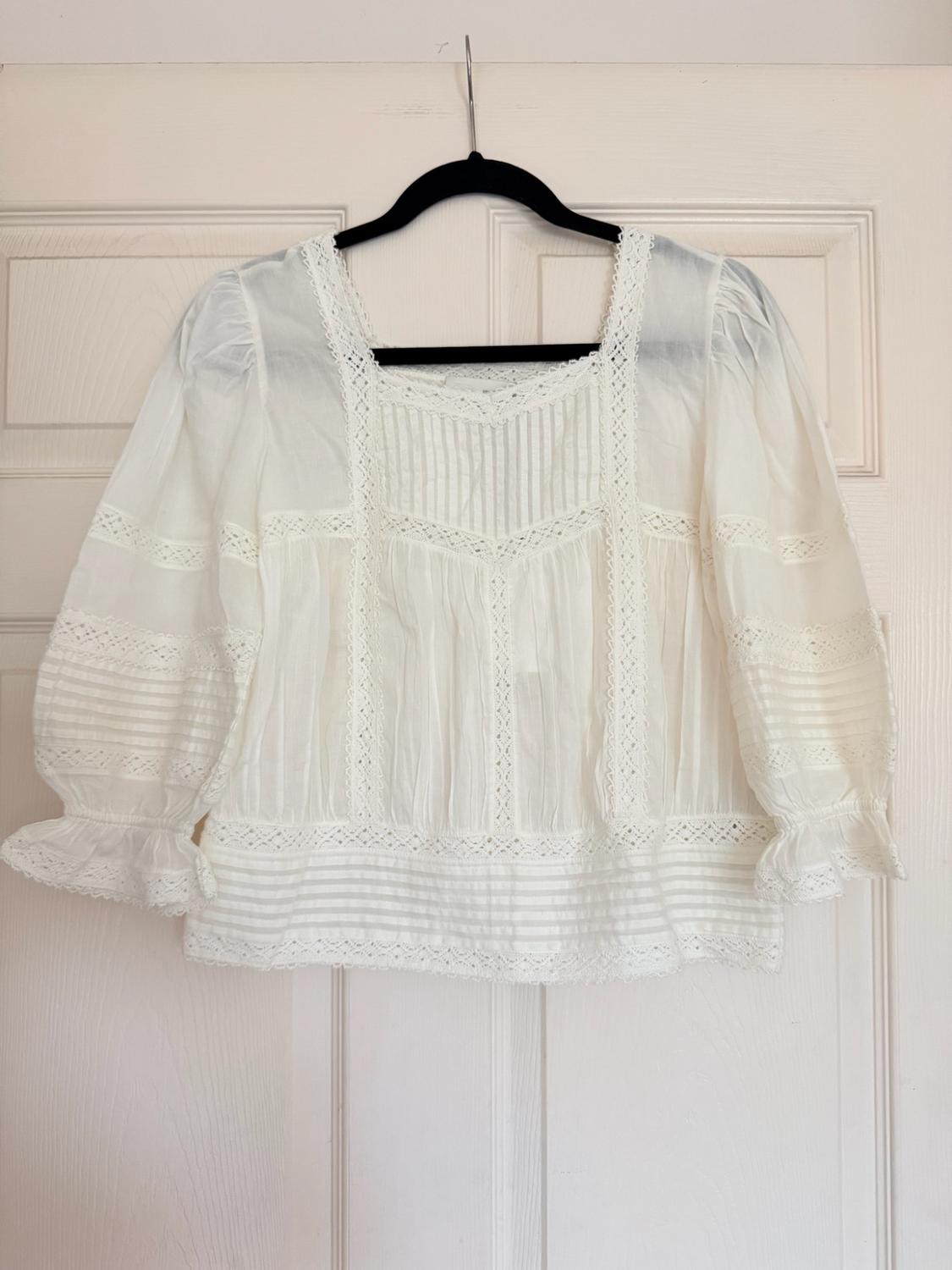 White blouse with a square neckline, lace, and ruffled sleeves, hanging on a black hanger on a white door.