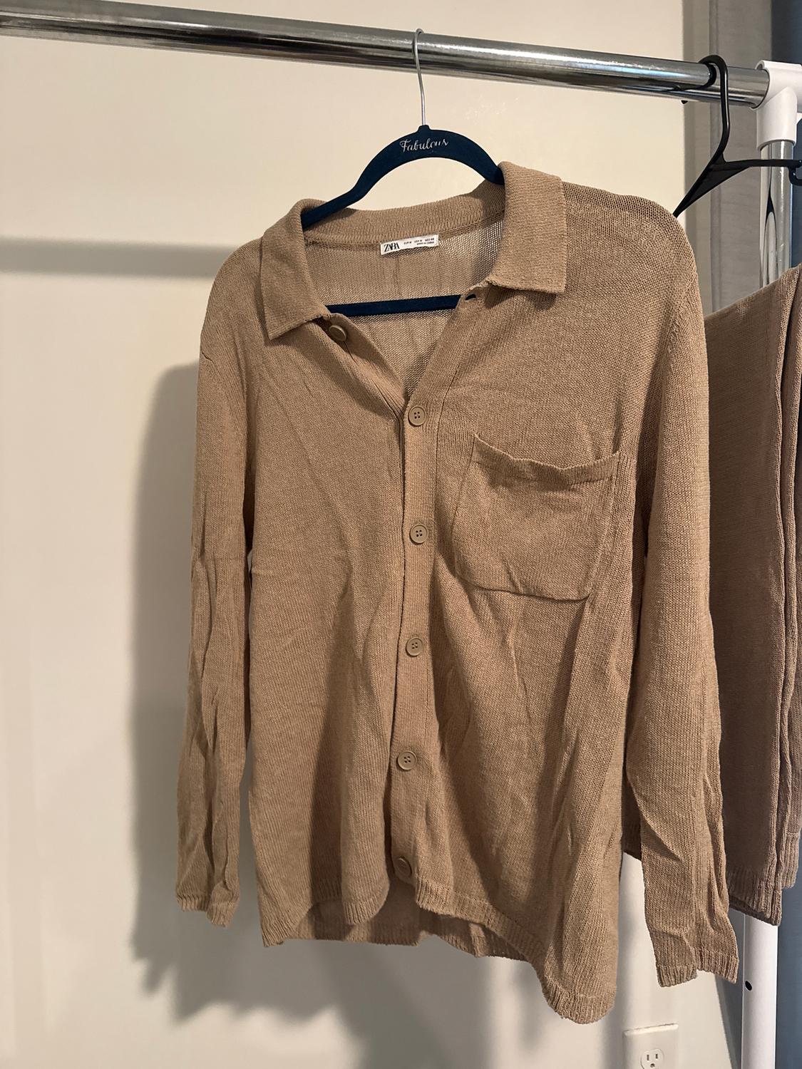 Light brown knitted shirt hanging on a blue hanger with the word "Fabulous". White background and visible metallic rail.