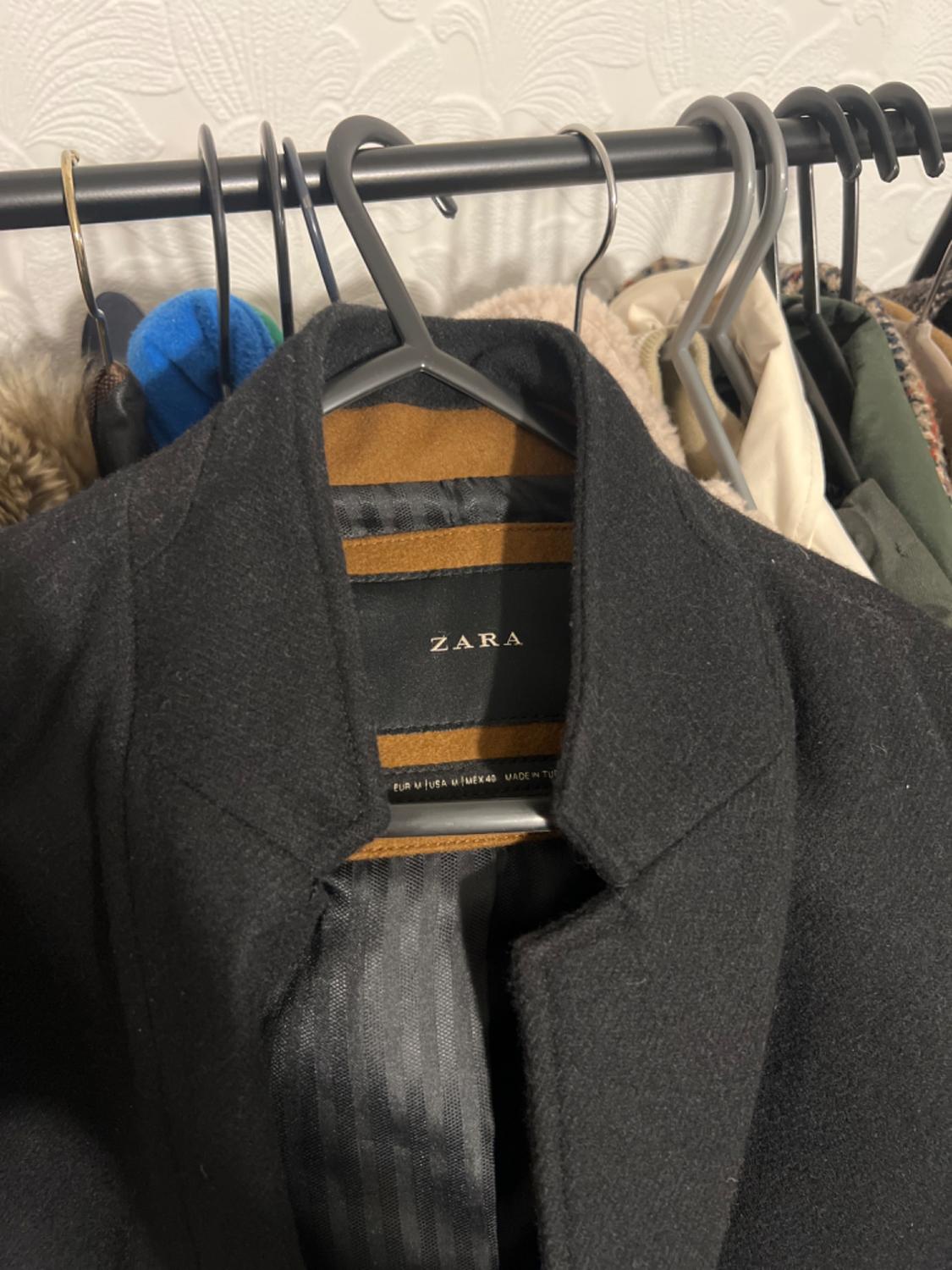 Black wool coat hanging on a clothes rail next to other garments. Visible label with the "ZARA" brand. White wallpaper background.