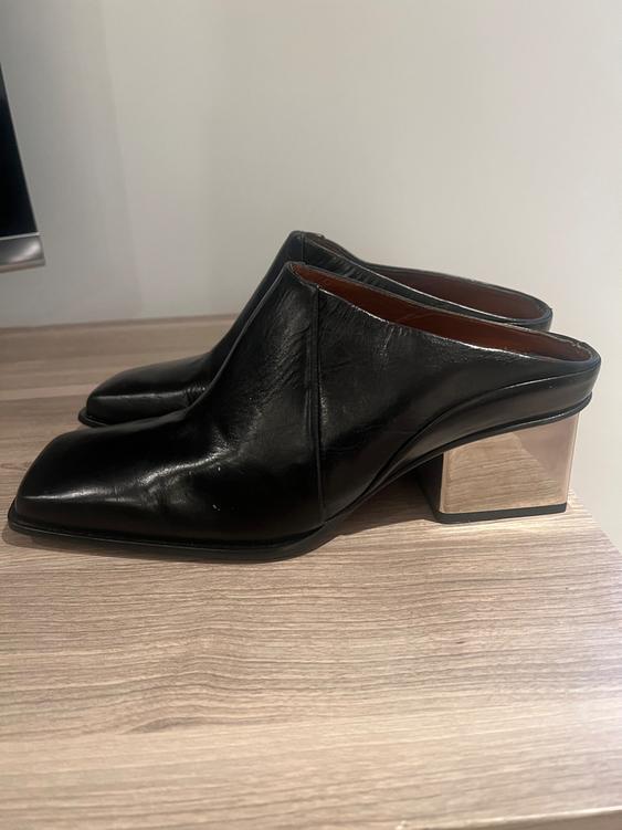 Black shoes with metallic block heels on a light wooden surface, with a white background.