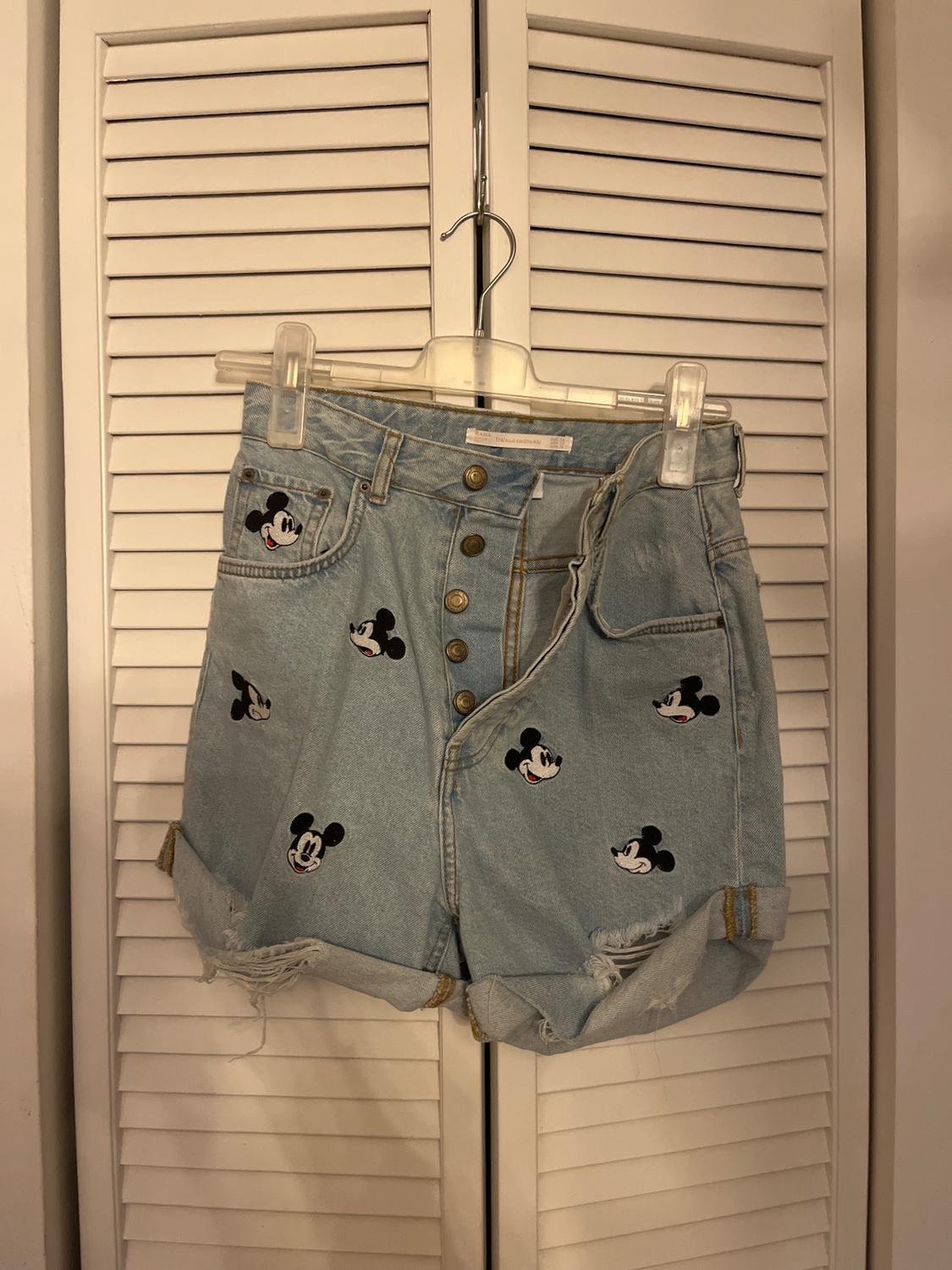 Light blue denim shorts with a Mickey Mouse faces print, hanging on a hanger in front of a white slatted door.