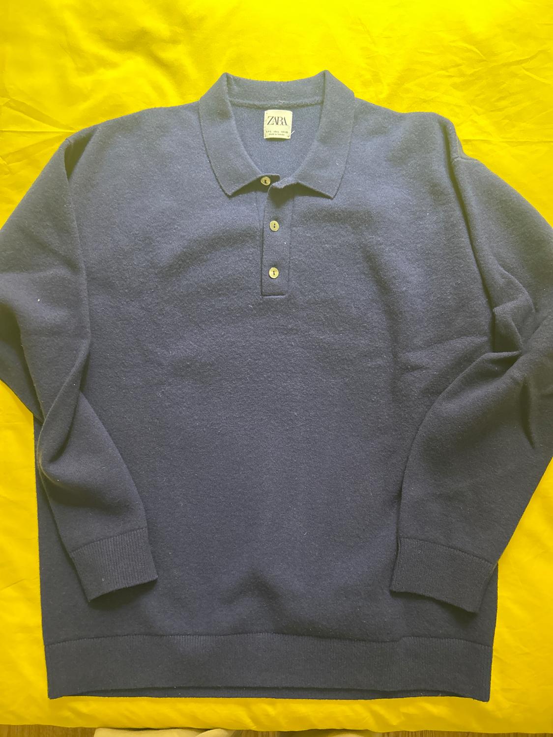 Long-sleeved blue polo shirt with a three-button placket, on a yellow background. Visible label on the collar.