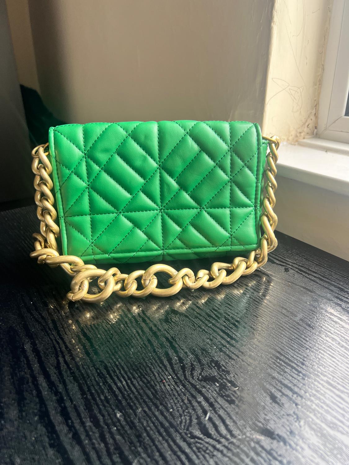 Green quilted handbag with a gold chain on a dark wooden table, next to a window lit by daylight.