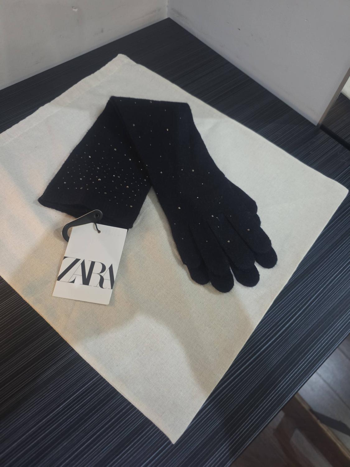 Long black gloves with small sparkles on a white fabric on a dark striped surface. Label with the text ZARA.