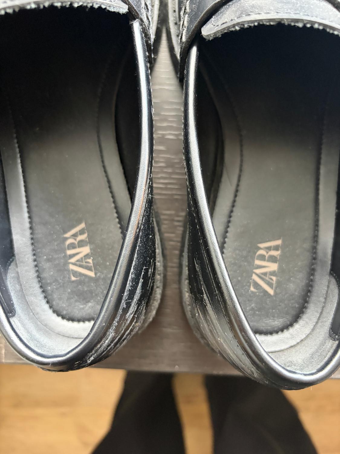 Black patent leather shoes with the "ZARA" brand visible on the insoles, viewed from above on a light wooden floor.