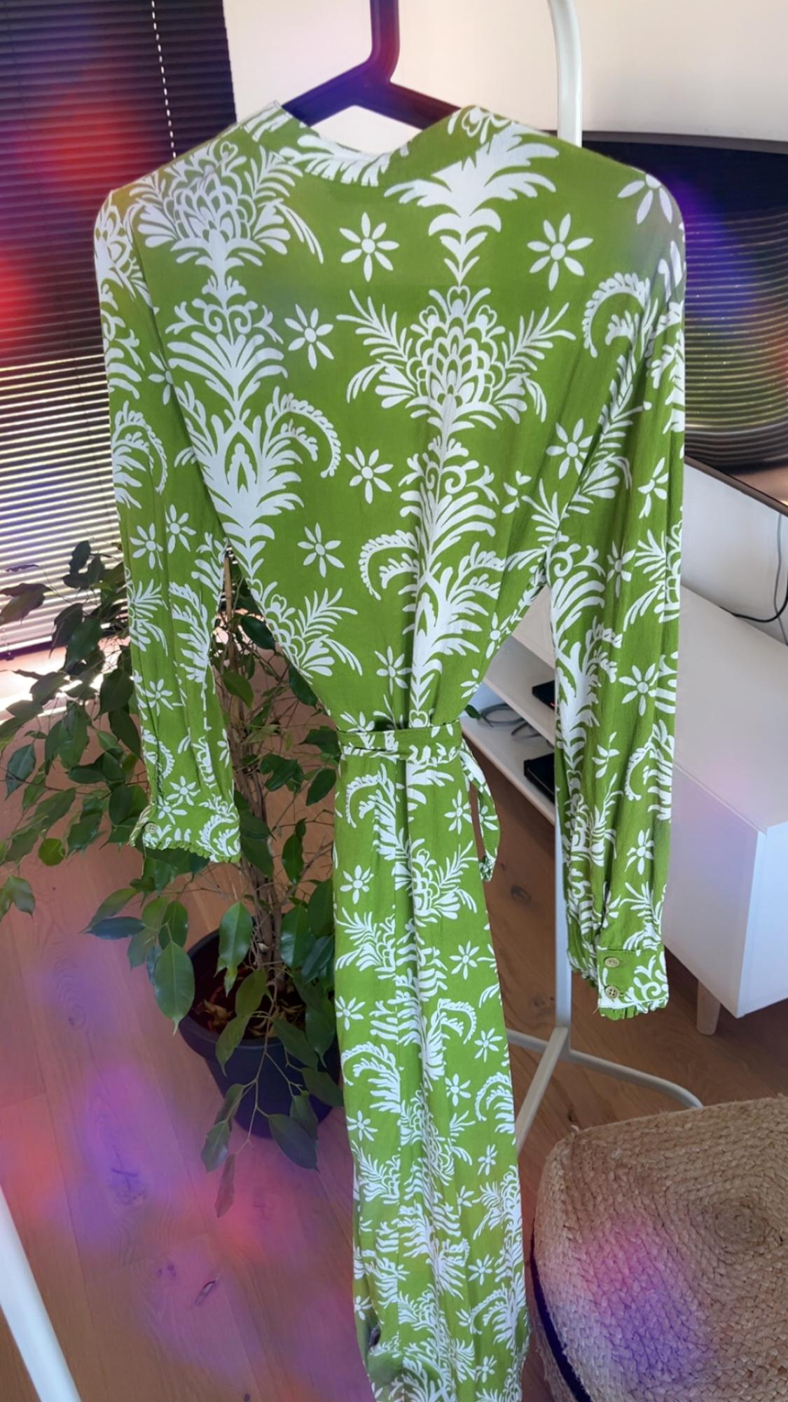 Green dress with white floral print hanging on a clothes rail. Background with plant and natural light coming through the window.