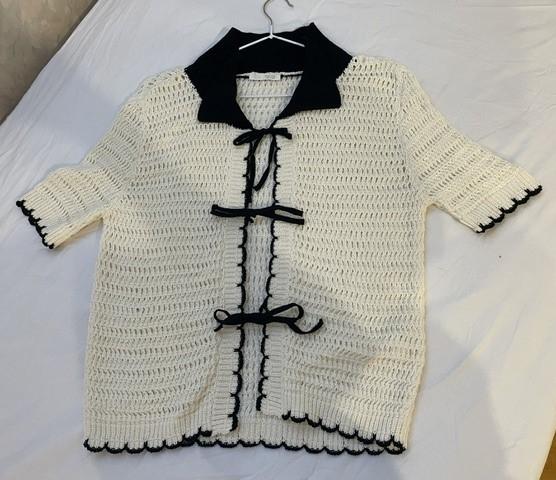White openwork knit top with a collar, front ties and black scalloped edges. Hanging on a hanger over a white sheet.