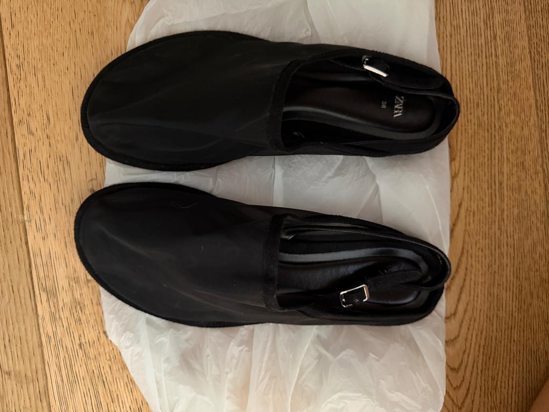 Black flat shoes with a strap and buckle on crumpled white paper on a wooden floor. Text: ZARA 38.