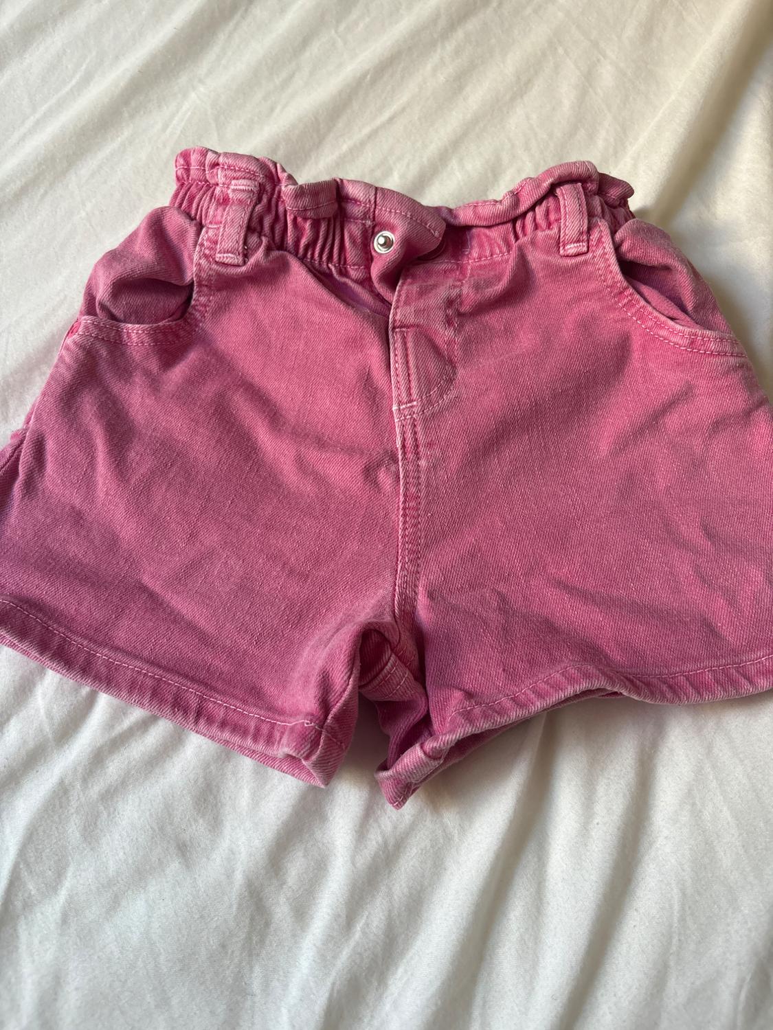 Pink denim shorts with an elasticated waistband, metal button and pockets, on crumpled white fabric. Casual style.