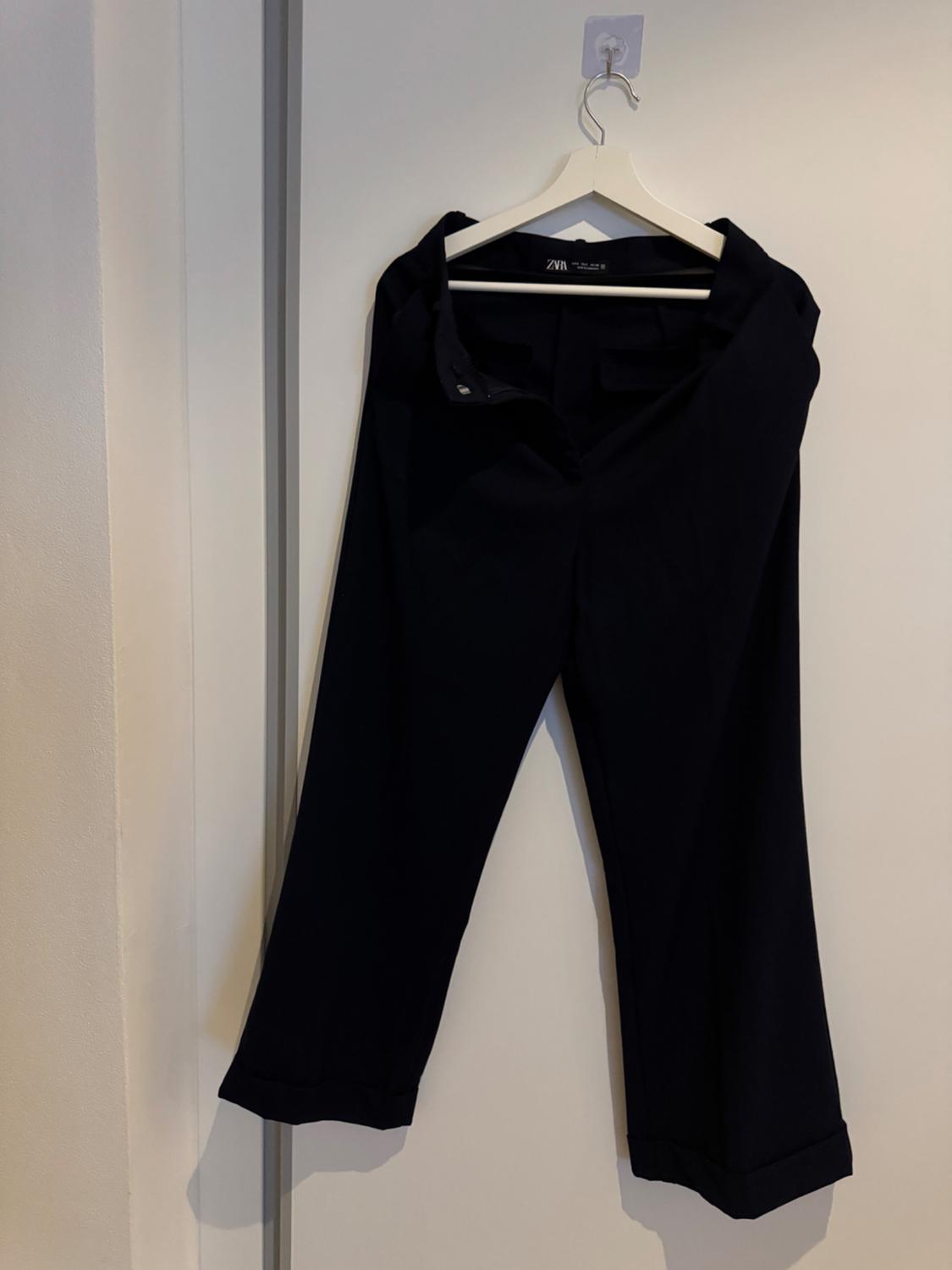 Front view of hanging black trousers with a button-fastening waistband with belt loops.