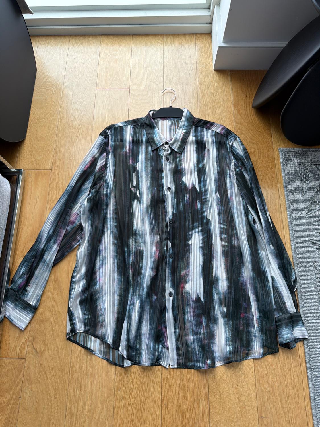Long-sleeved shirt with an abstract print in dark and light tones, hanging on a hanger over a light wooden floor.