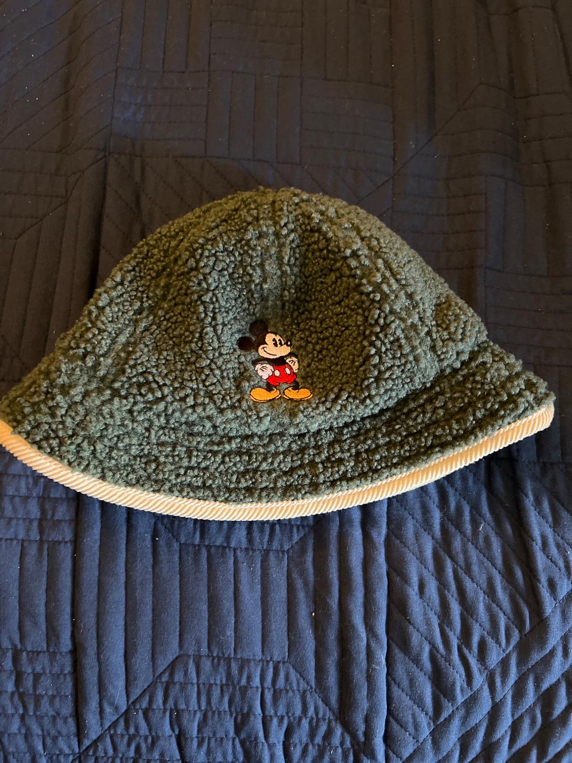 Green woolly hat with an embroidered cartoon character in the centre, on a blue quilt with a geometric pattern.