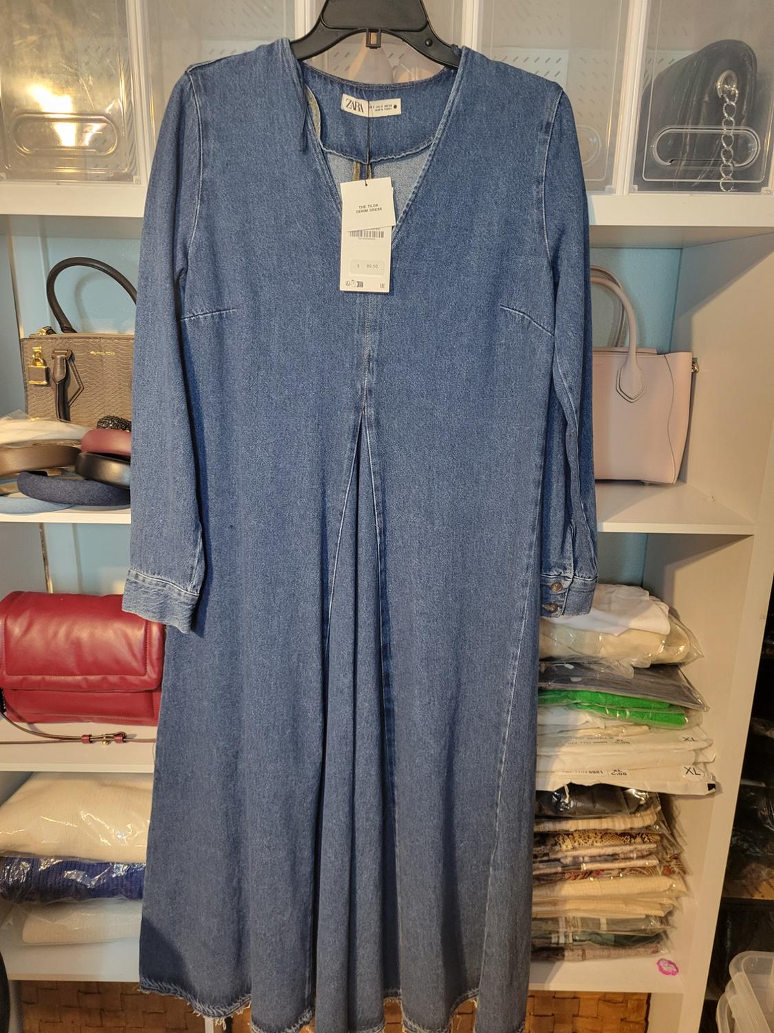 Long blue denim dress hanging on a hanger, with visible tag. Background with shelves and multi-colored handbags.