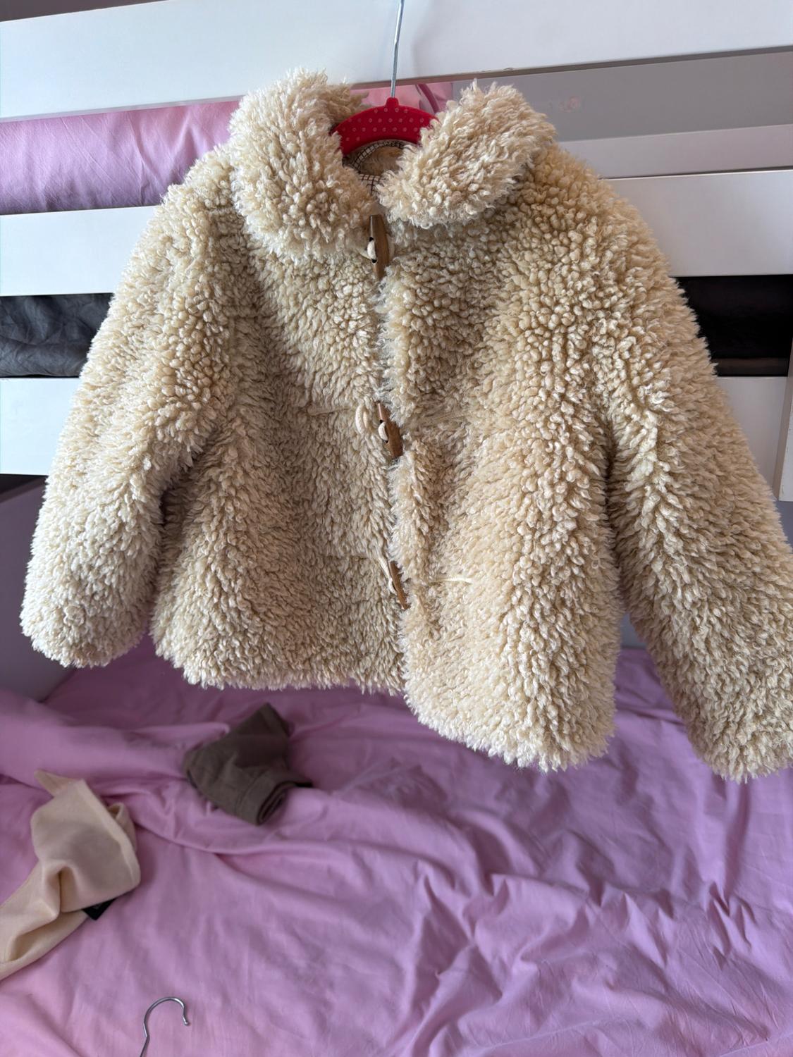 Beige fluffy-textured coat with buttons, hanging on a pink hanger over a white bed with pink sheets.