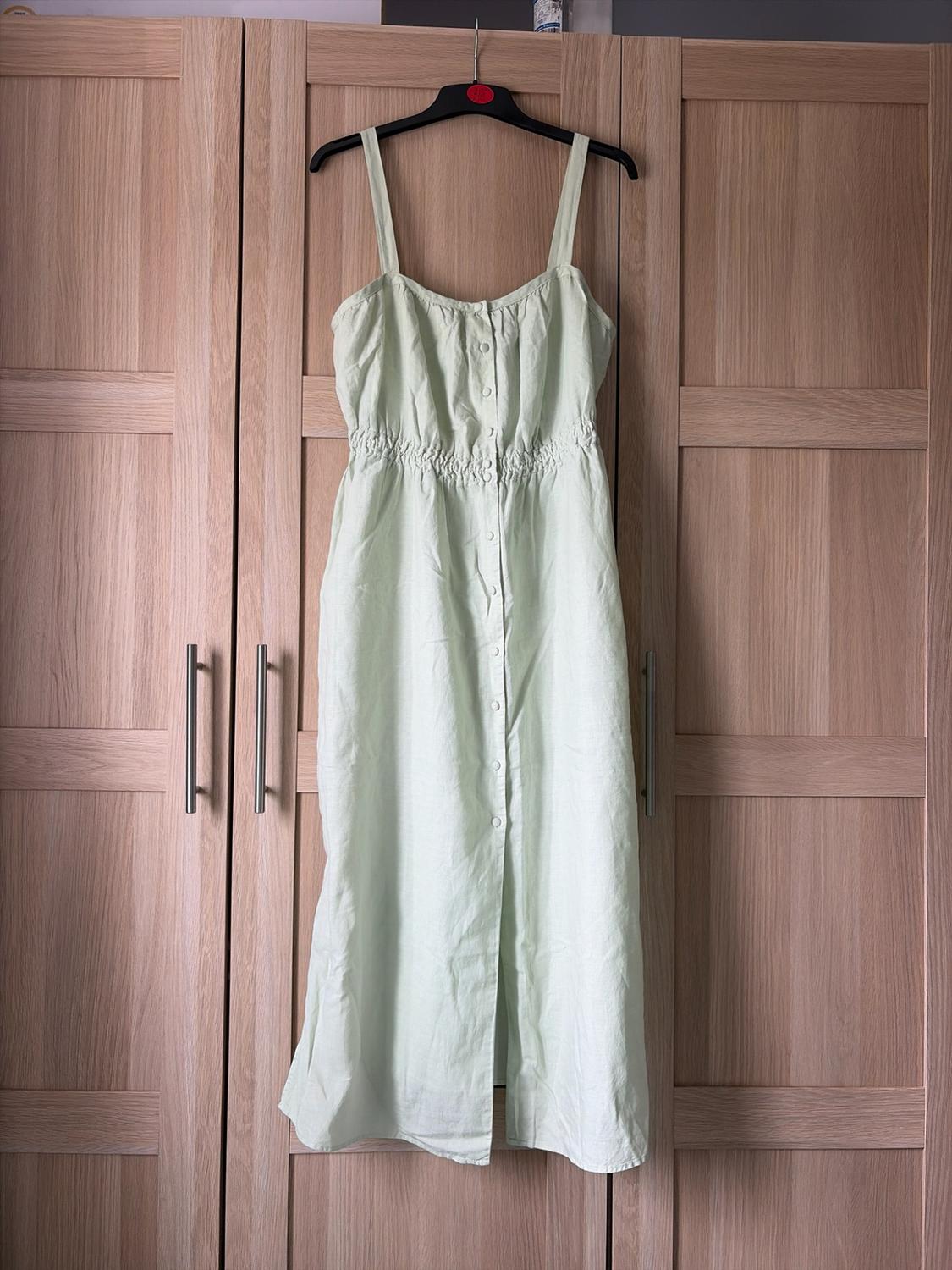 Light green camisole dress, with front buttons and an elasticated waist, hanging on a black hanger in front of a light wood wardrobe.
