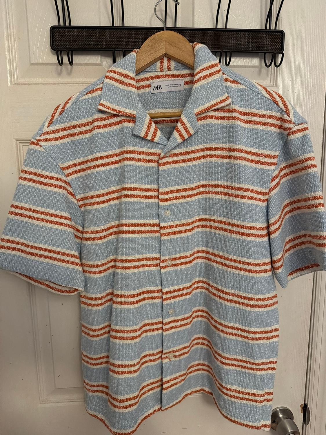 Light blue, white, and orange horizontal striped shirt, hanging on a wooden hanger in front of a white door.