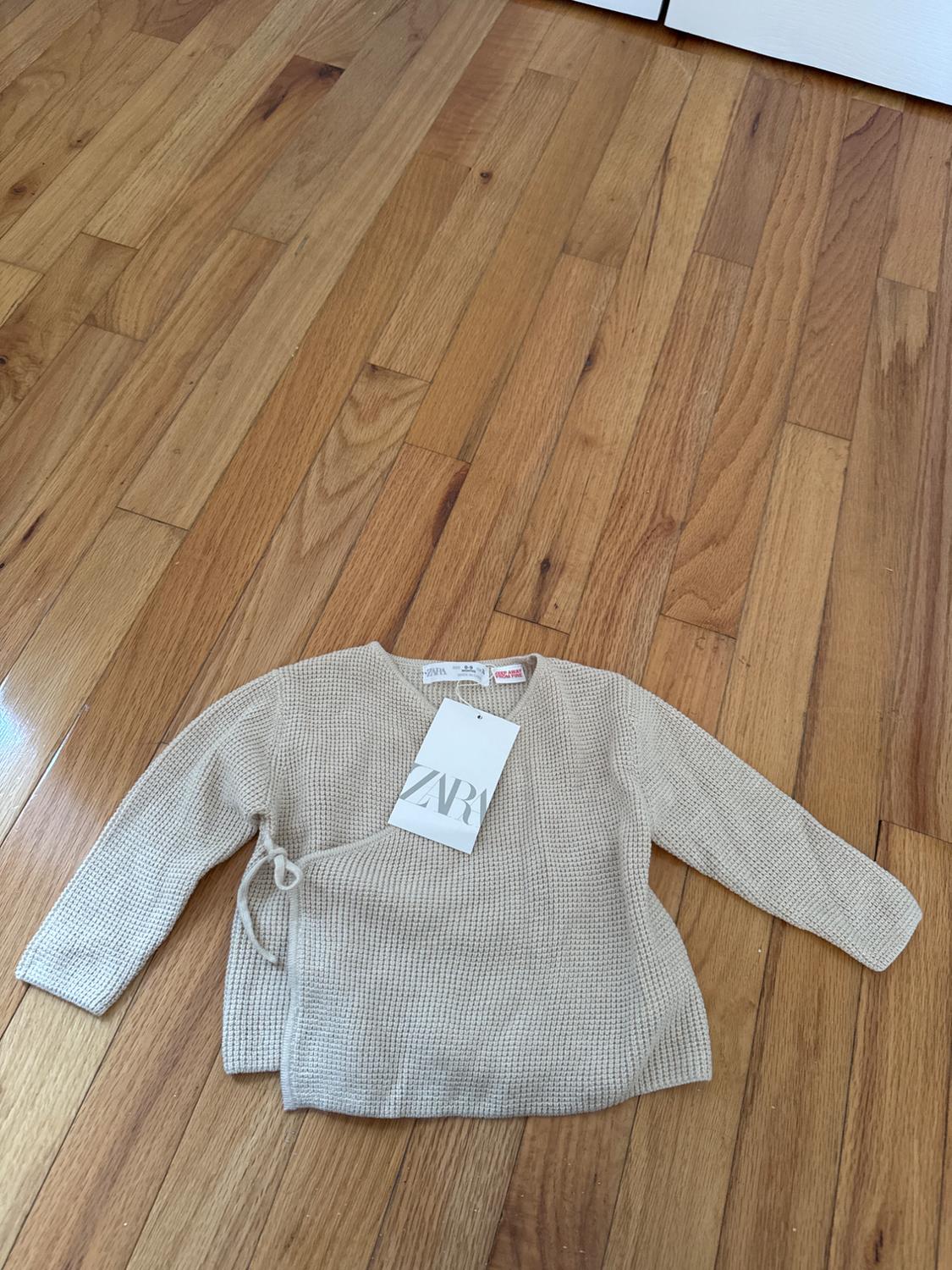 Beige fine knit jumper with ZARA label on light wood floor. Long sleeves and side tie detail.
