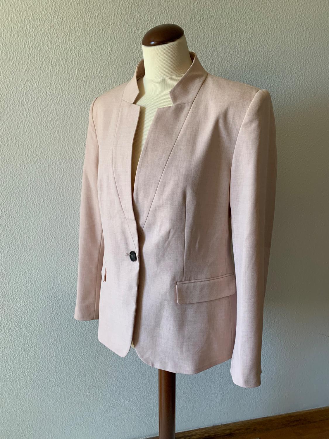 Mannequin in a beige single-button jacket with an open collar and front pockets, against a white wall background.
