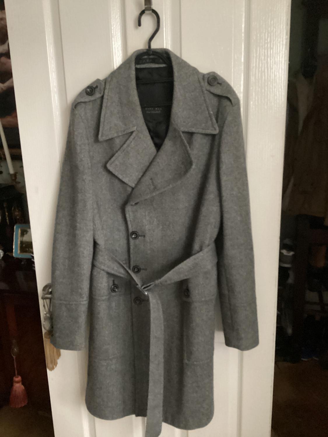 Grey coat with a belt and buttons, hanging on a white door. Interior setting with decorative details in the background.