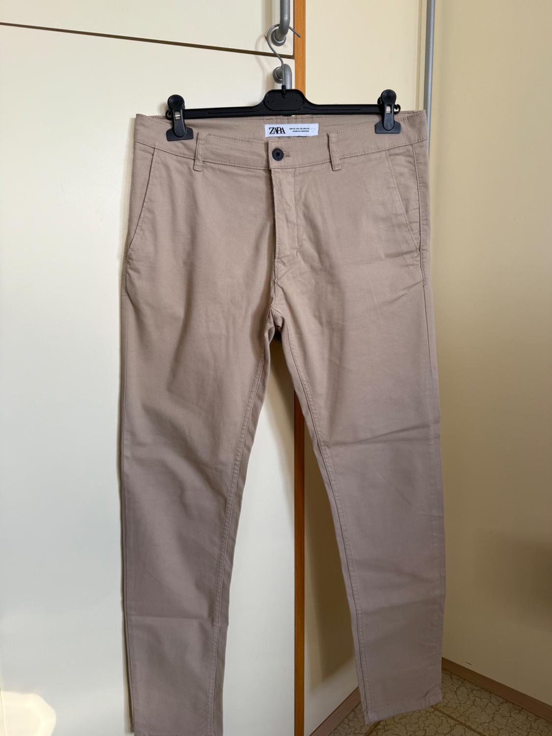 Beige trousers hanging on a black hanger with clips, visible Zara label, white door and beige wall background.