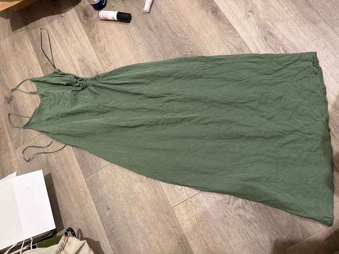 Olive green long dress with thin straps on a wooden floor. Around it are beauty products and a paper bag.