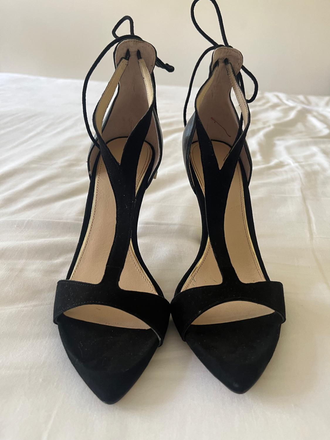 Black high-heeled T-bar sandals with ankle straps, set on a crumpled white fabric surface.