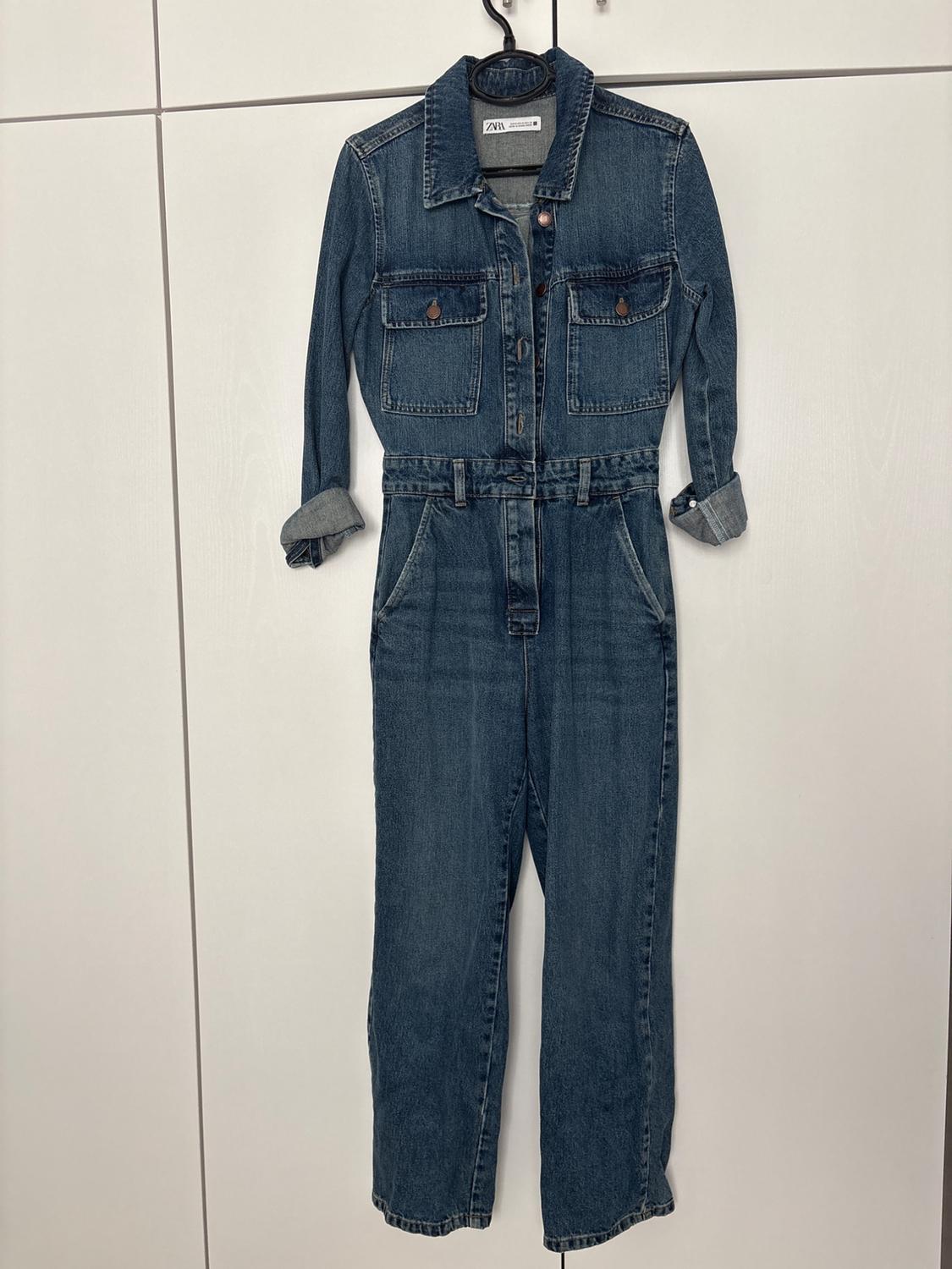Blue denim jumpsuit with rolled-up sleeves and pockets, hanging on a black hanger in front of a white wardrobe. Label with the text ZARA.
