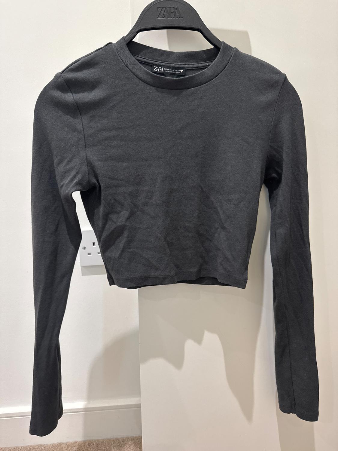 Black long-sleeved T-shirt hanging on a hanger with visible branding. White background and beige carpeted floor.