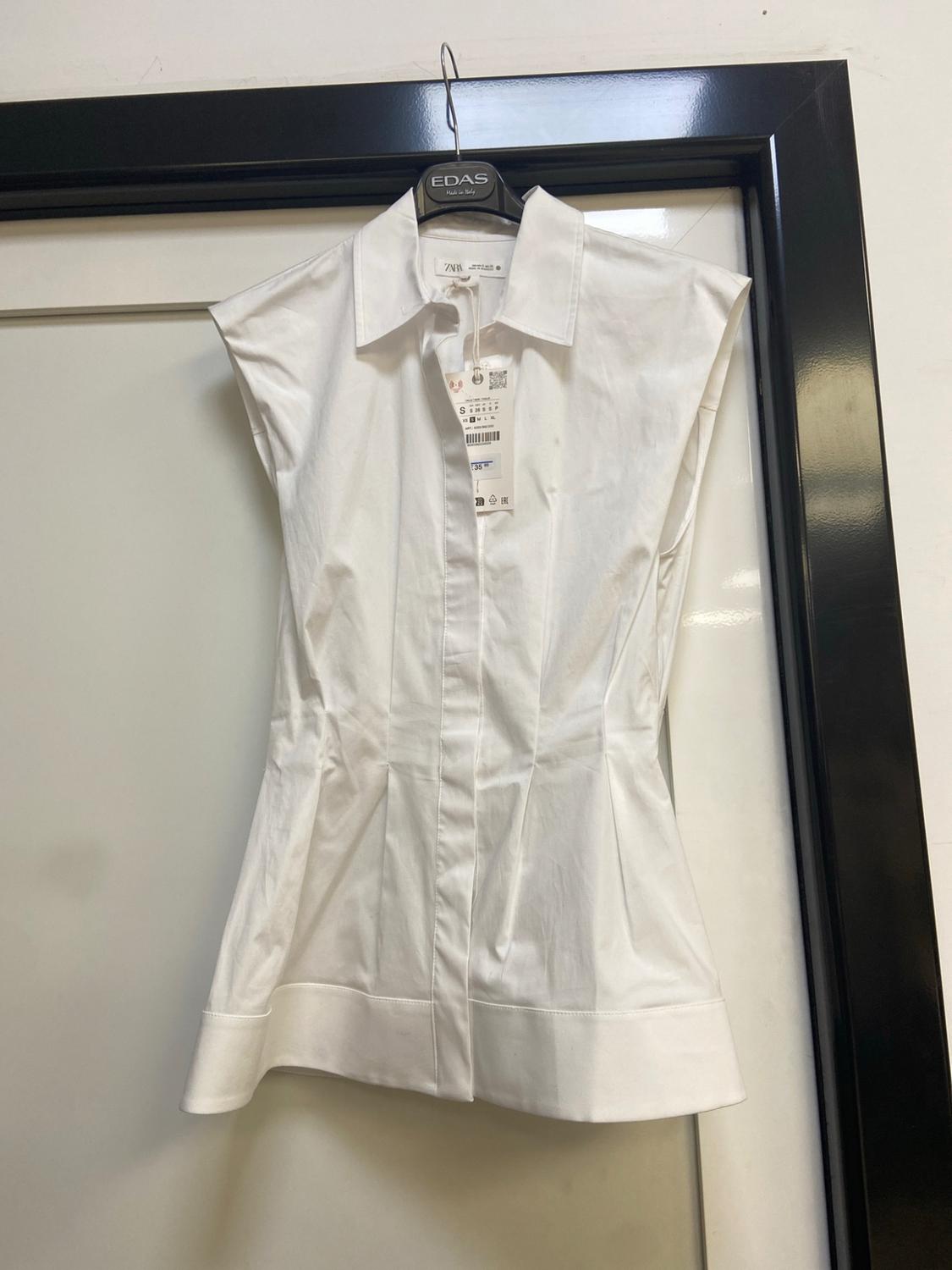 Fitted sleeveless white shirt hanging on a door. Hanger with the text EDAS Made in Italy and a ZARA S label.