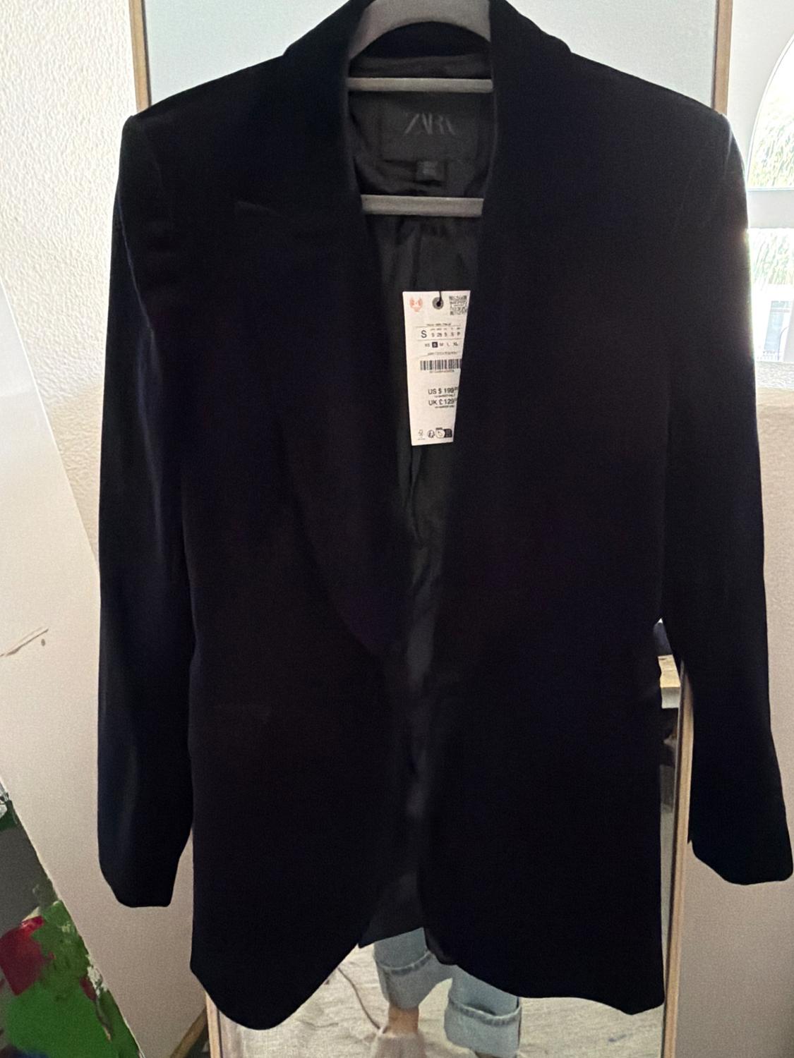 Black jacket hanging on a hanger, with a visible tag. Reflection of a person in jeans in the mirror behind.