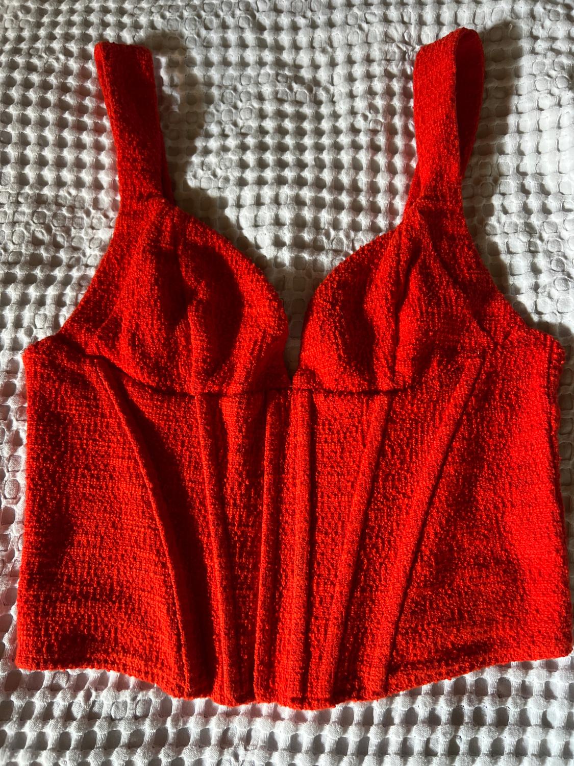 Red textured corset-style top with wide straps, on a white embossed fabric. Relaxed composition with sharp shadows.