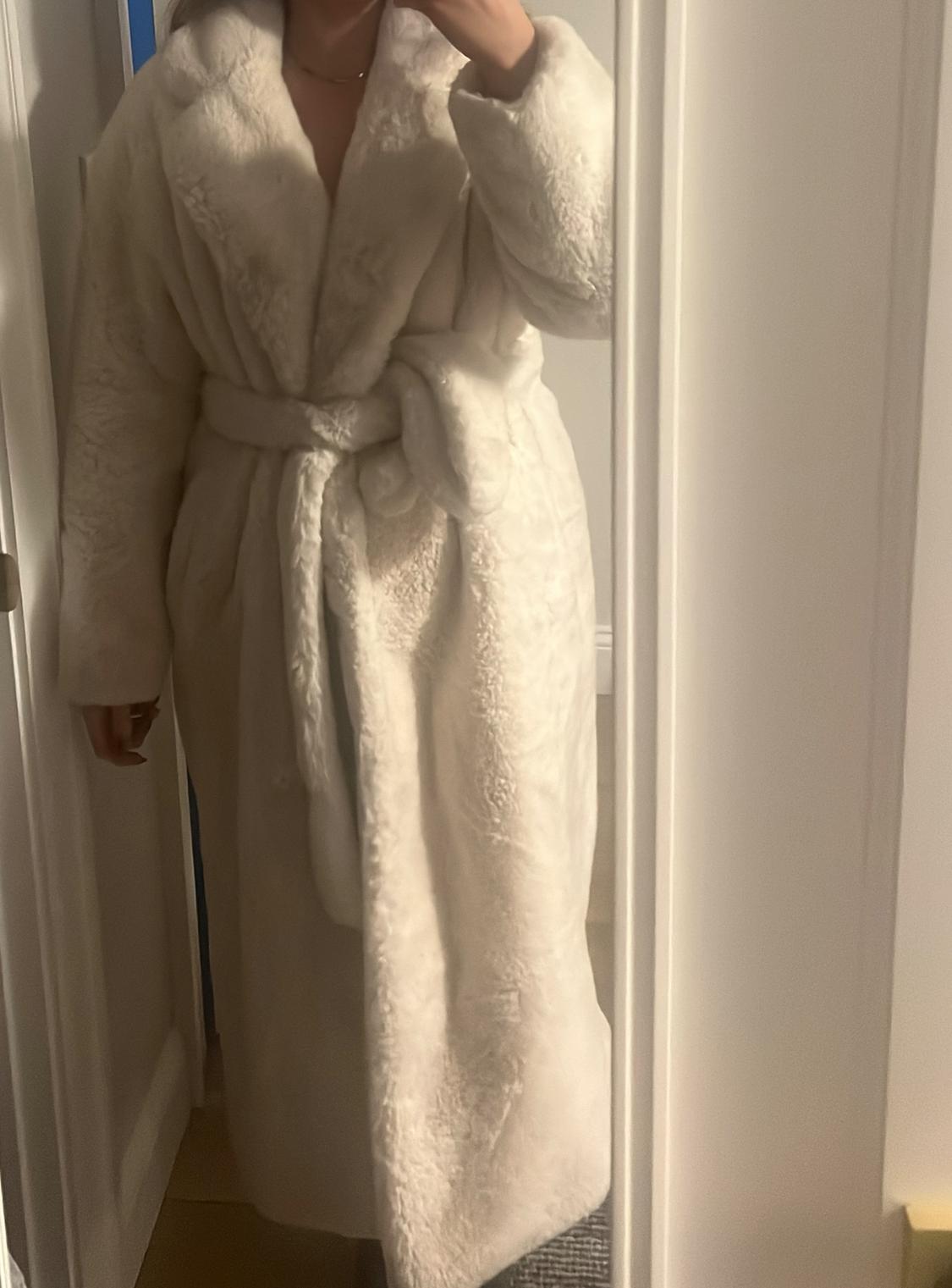Person in a long white faux fur coat, tied with a belt, posing in front of a mirror in a well-lit indoor environment.
