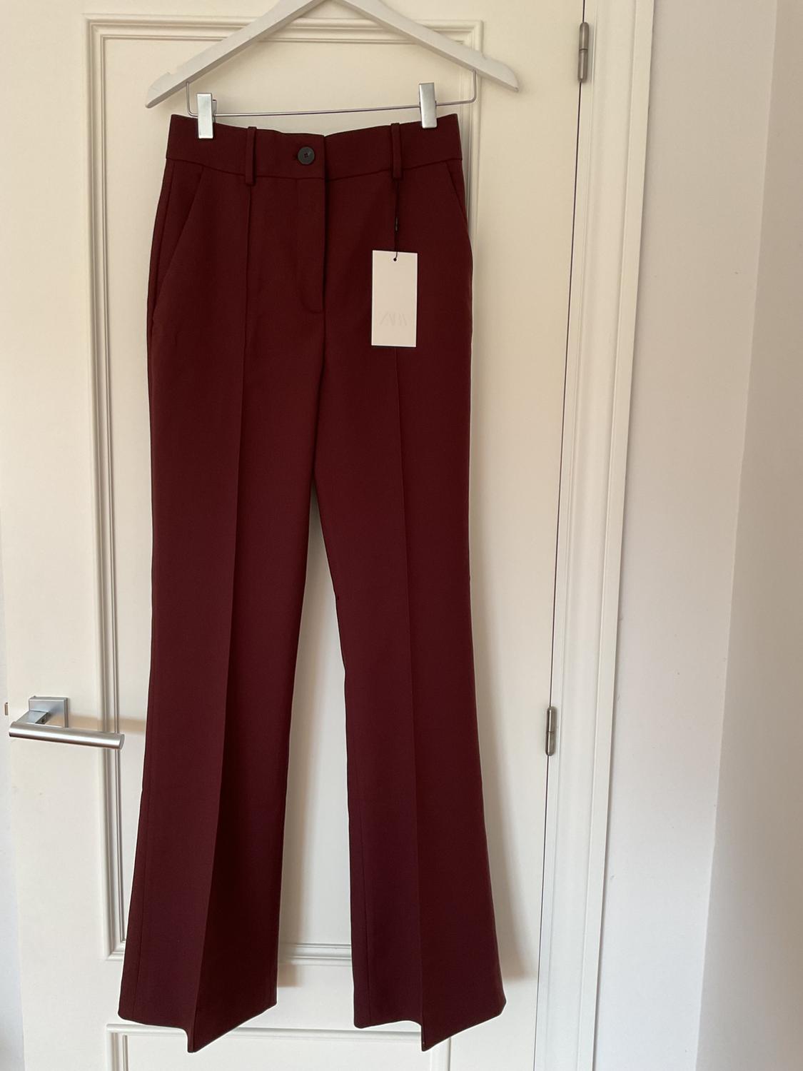 Burgundy wide-leg trousers hanging on a white hanger over a white door, with visible tag.