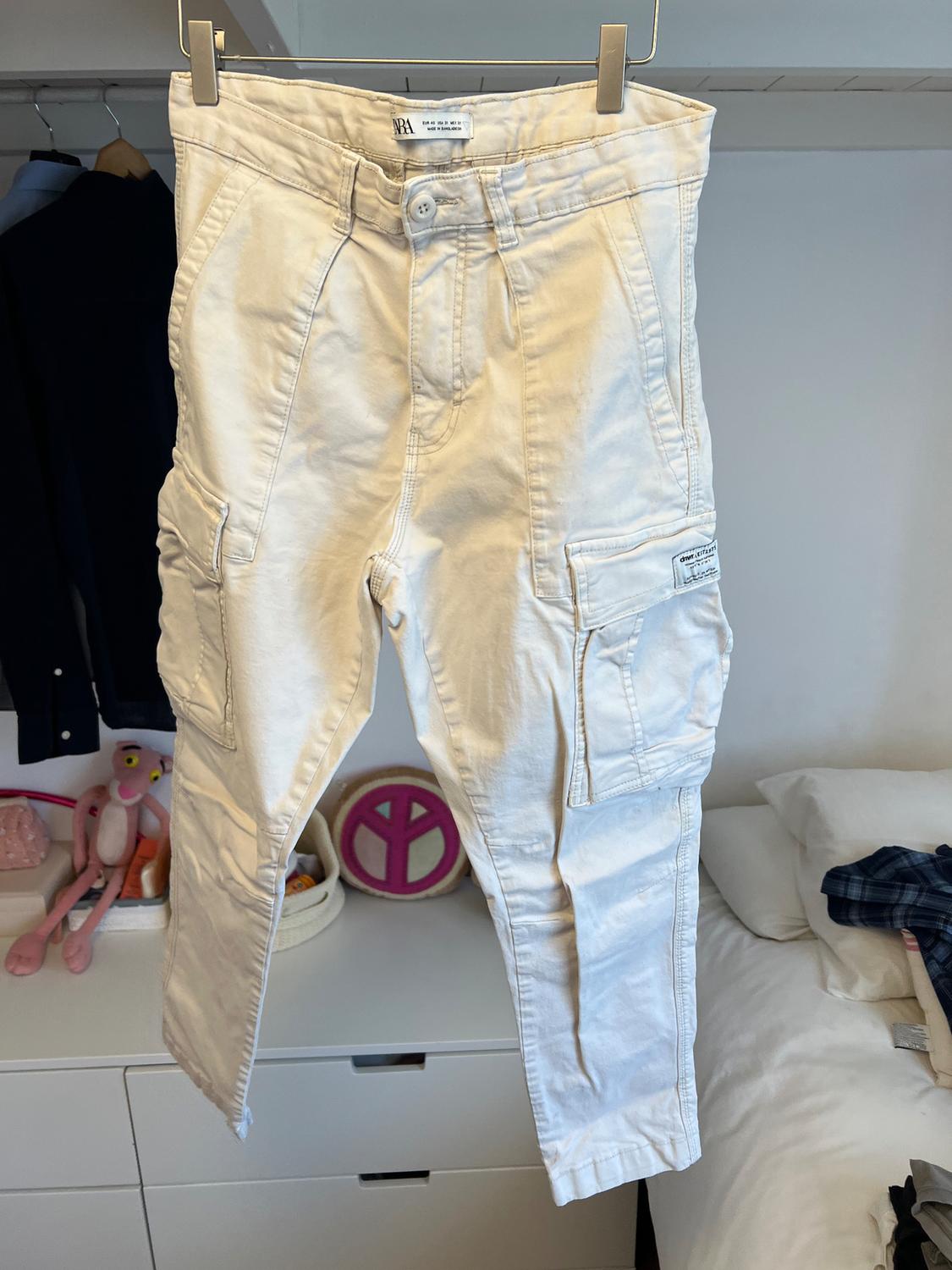 White cargo trousers hanging on a hanger over a bed. Background with toys and clothes in an open wardrobe.